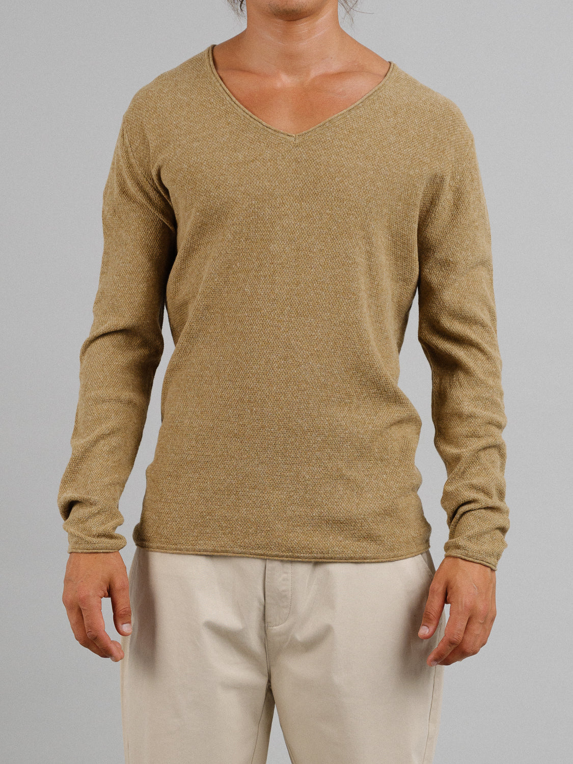 A person models the Amalfi Deep V Knitted Top by ID Menswear in tan, paired with beige pants against a gray backdrop. Their head isnt visible, highlighting the deep V-neck design and the smooth linen-cotton blend of this refined ensemble.