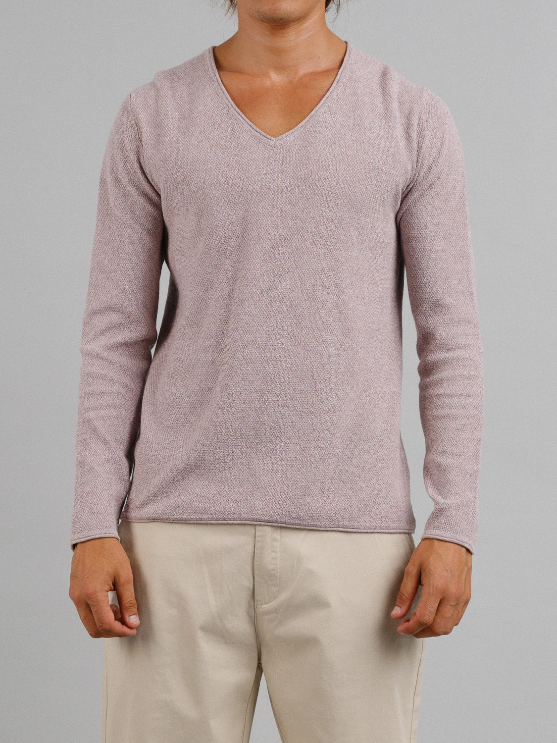 Someone models an Amalfi Deep V Knitted Top by ID Menswear, featuring a light pink shade and long sleeves, perfectly paired with beige pants against a gray background. The focus is on the torso, highlighting the elegant linen-cotton blend.
