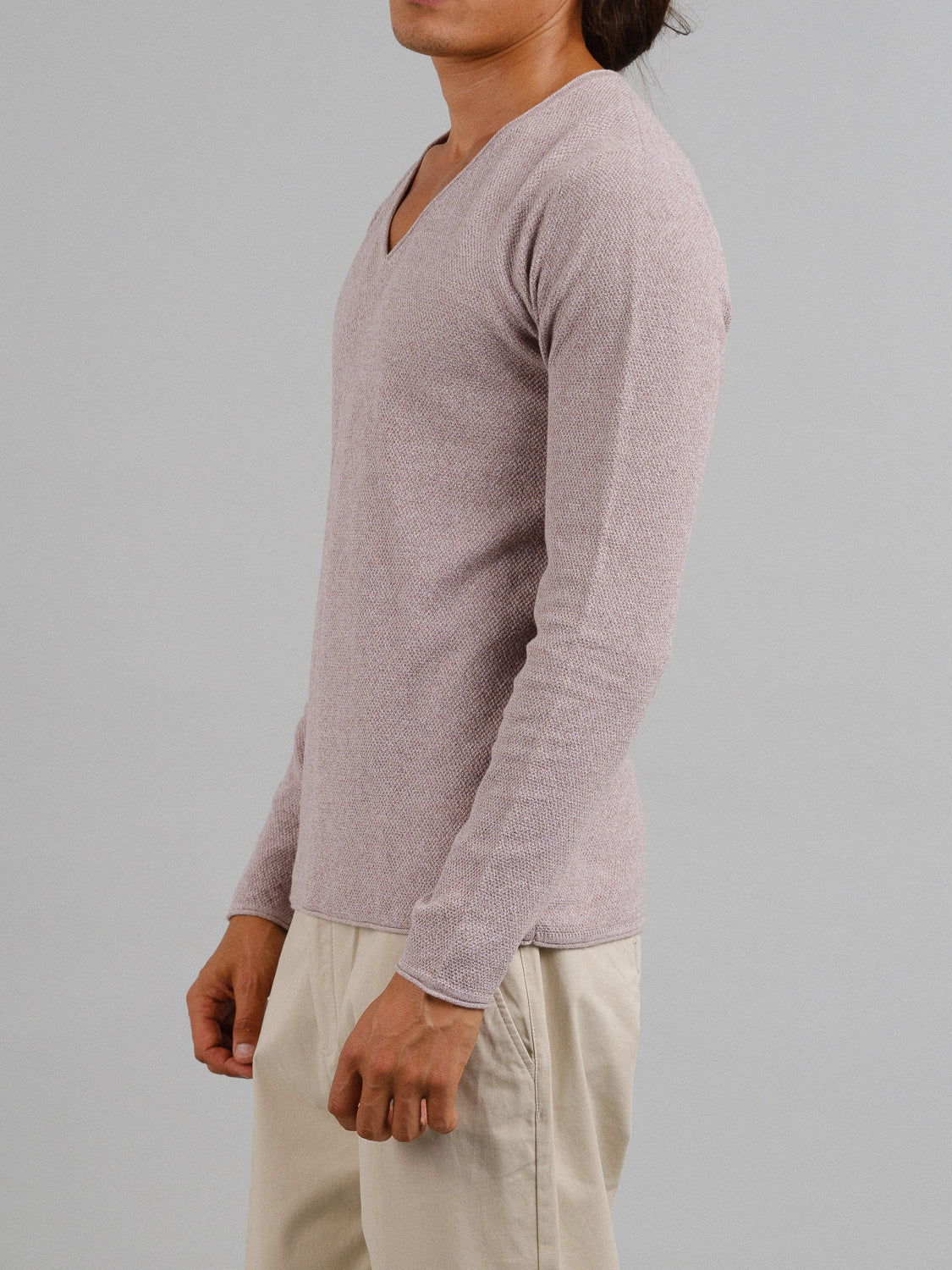 A person stands sideways to the left against a gray background, relaxed arms down, wearing a light pink Amalfi Deep V Knitted Top from ID Menswear and beige pants.