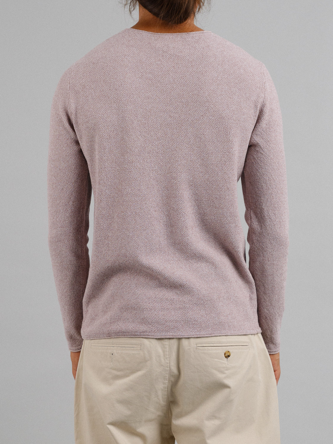 Rear view of a person wearing a light pink Amalfi Deep V Knitted Top by ID Menswear and beige pants. The linen and cotton blend adds texture. The individual has brown hair tied back against a plain gray backdrop.