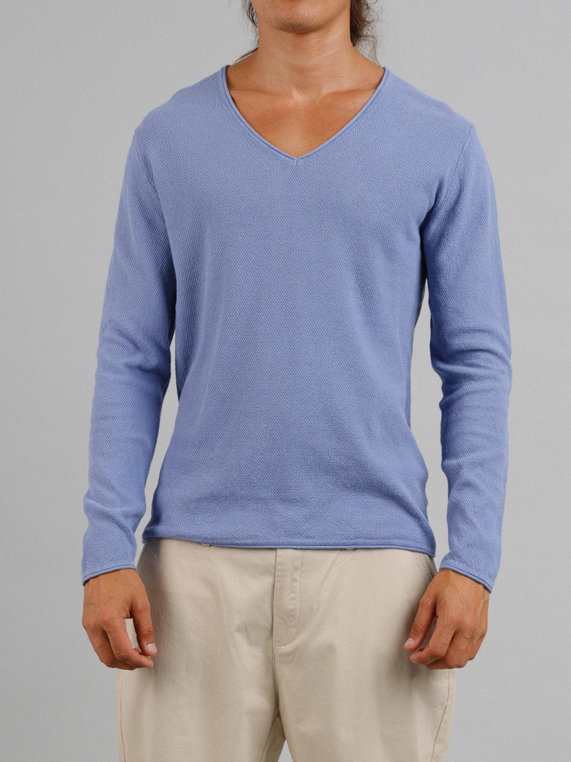 A person models a light blue Amalfi Deep V Knitted Top from ID Menswear paired with beige pants, set against a gray backdrop, showcasing the upper body and clothing details.