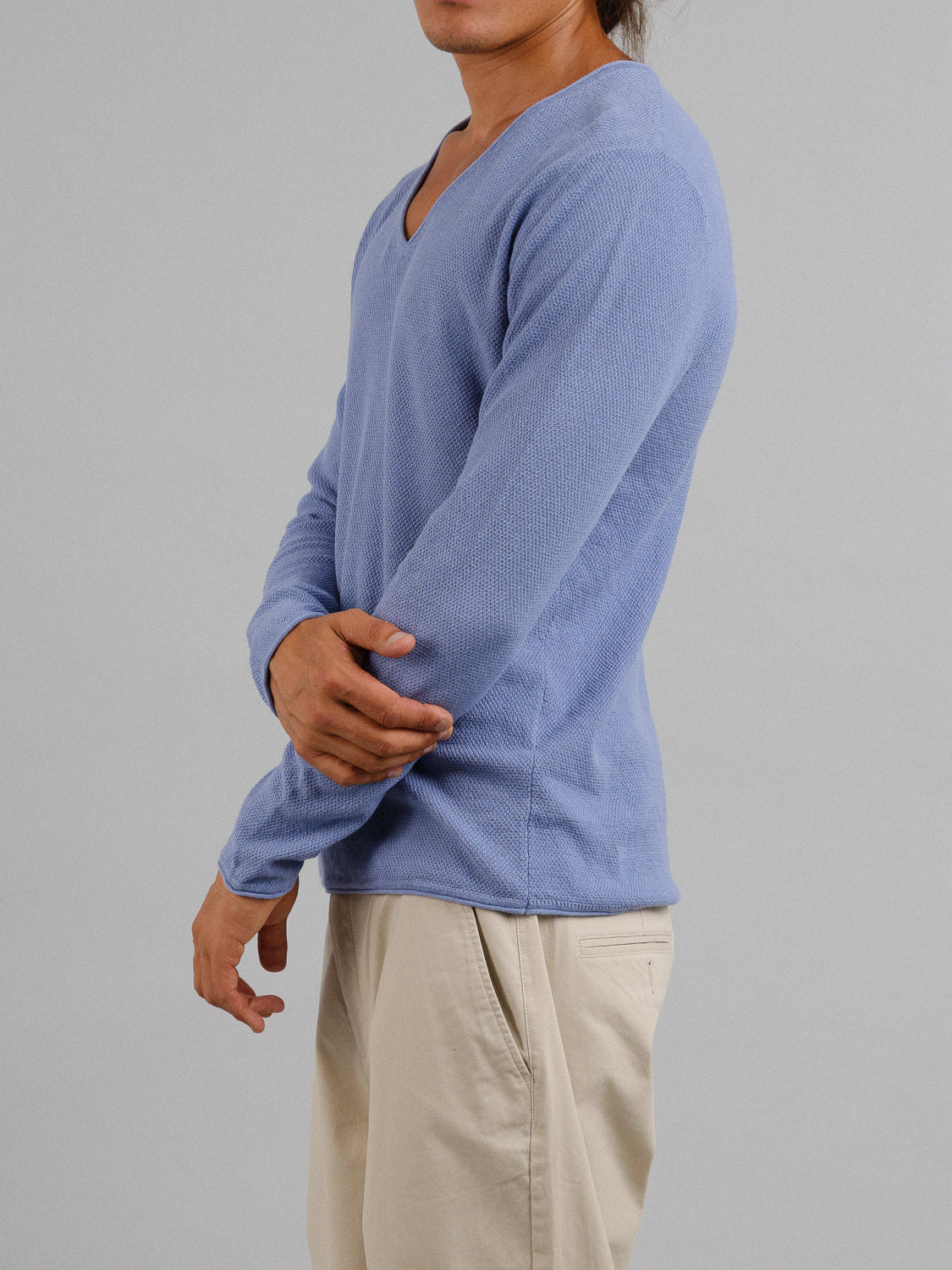 Against a plain gray background, someone with arms crossed wears the Amalfi Deep V Knitted Top by ID Menswear in a light blue linen-cotton blend and beige pants. Their face isnt visible.