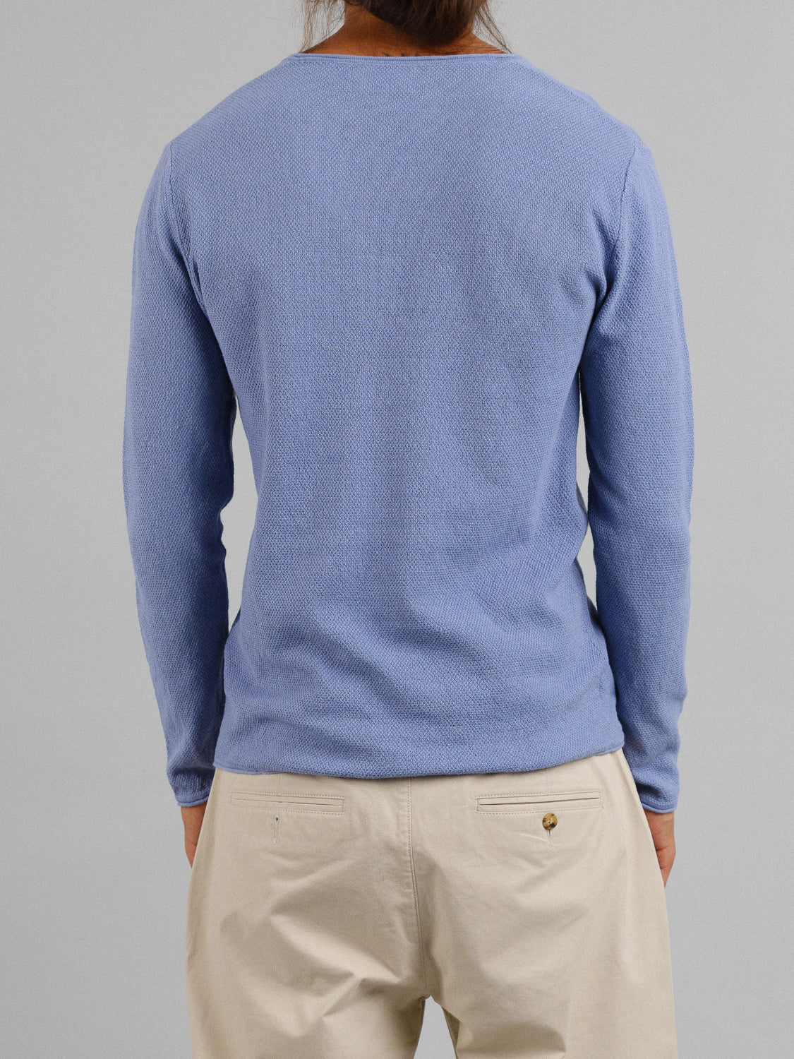 A person is seen from behind wearing a light blue Amalfi Deep V Knitted Top by ID Menswear and beige pants, against a gray background. The linen-cotton blend drapes subtly as they face away, hands in pockets.