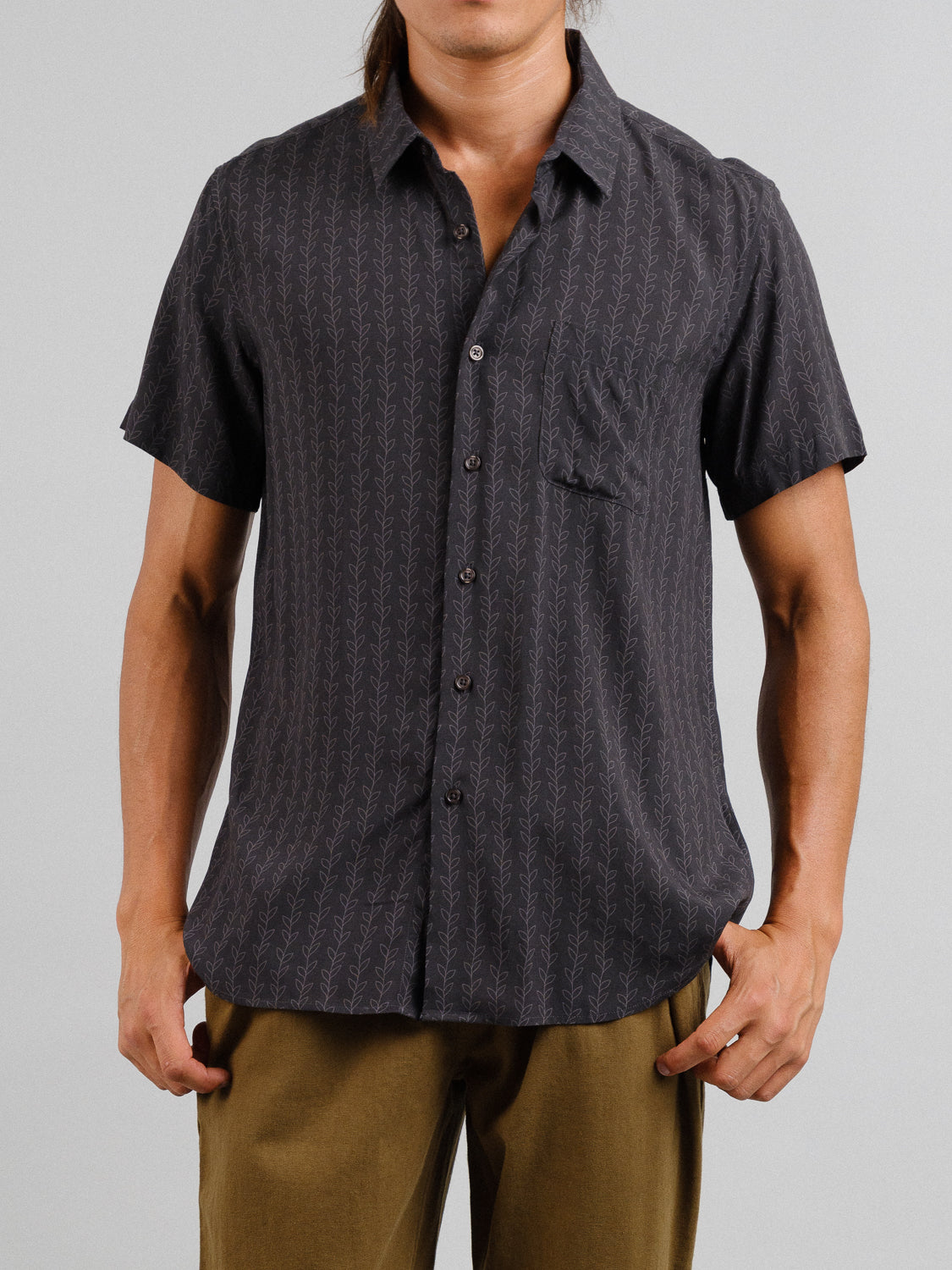 Against a plain backdrop, an individual wears the Kelp Short Sleeve Printed Rayon Shirt by ID Menswear, featuring vibrant prints and a subtle leaf-like design. Paired with olive green pants, the shirt includes a convenient chest pocket for effortless style.