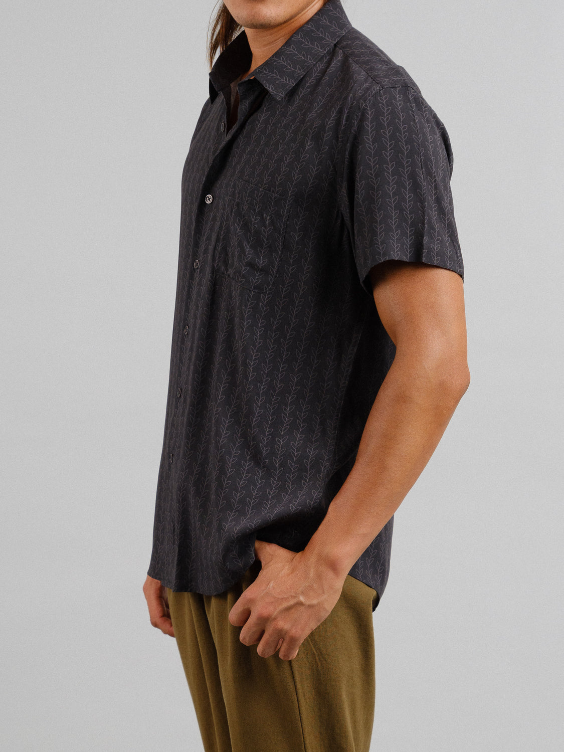 A person is standing against a neutral background, wearing the Kelp Short Sleeve Printed Rayon Shirt by ID Menswear with subtle patterns and light brown pants. The shirt features a collar and buttons down the front. Only their upper half is visible.