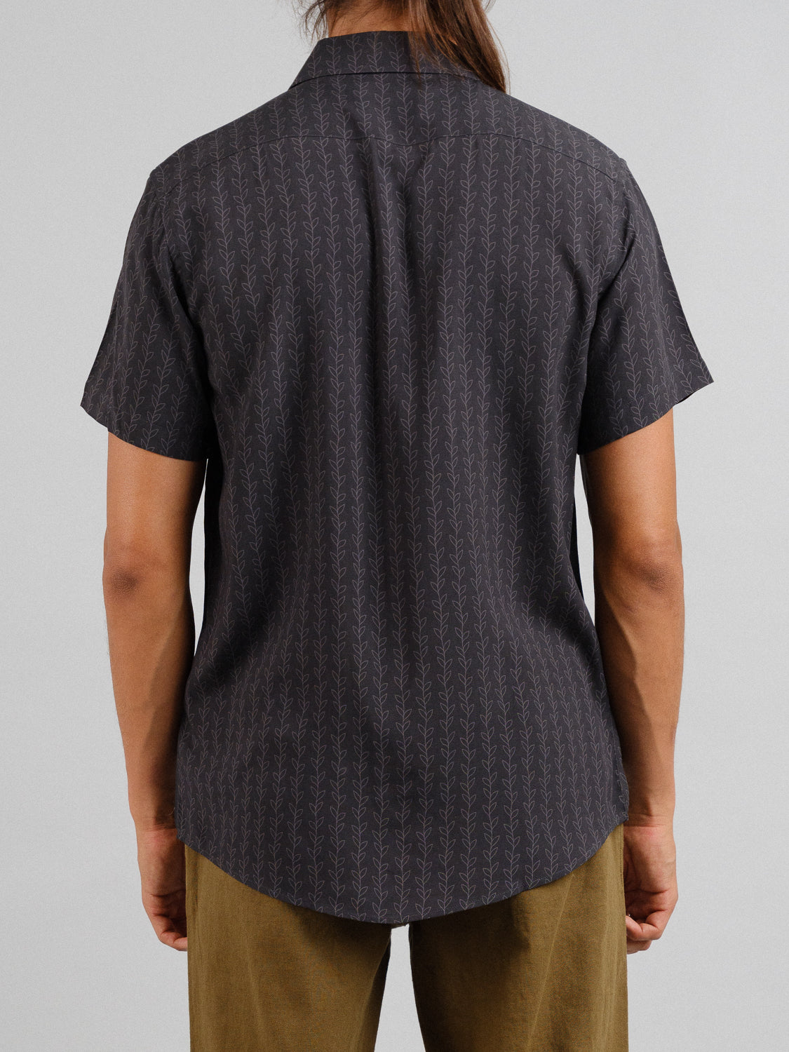 A person is seen from behind in an ID Menswear Kelp Short Sleeve Printed Rayon Shirt paired with olive green pants. Their long brown hair flows down, and the plain gray backdrop enhances the shirts vibrant patterns.
