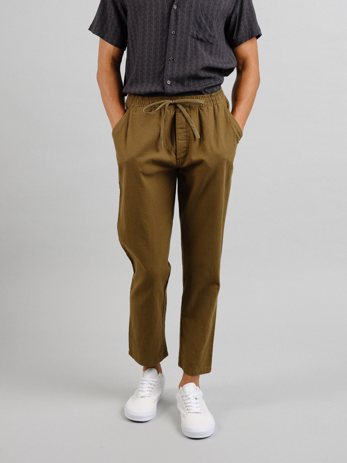 A person in ID Menswears Laguna Linen Cotton Drawstring Pant with a relaxed crop fit and brown color paired with a dark gray short-sleeve shirt stands in a neutral pose. Their hands are casually tucked into pockets, complemented by white sneakers on a plain light gray background.