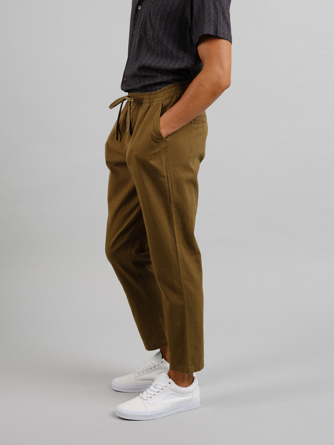 Wearing ID Menswears Laguna Linen Cotton Drawstring Pant in a relaxed crop fit, paired with a dark shirt and white sneakers, a person stands against a plain gray backdrop, exuding casual style with hands in pockets.