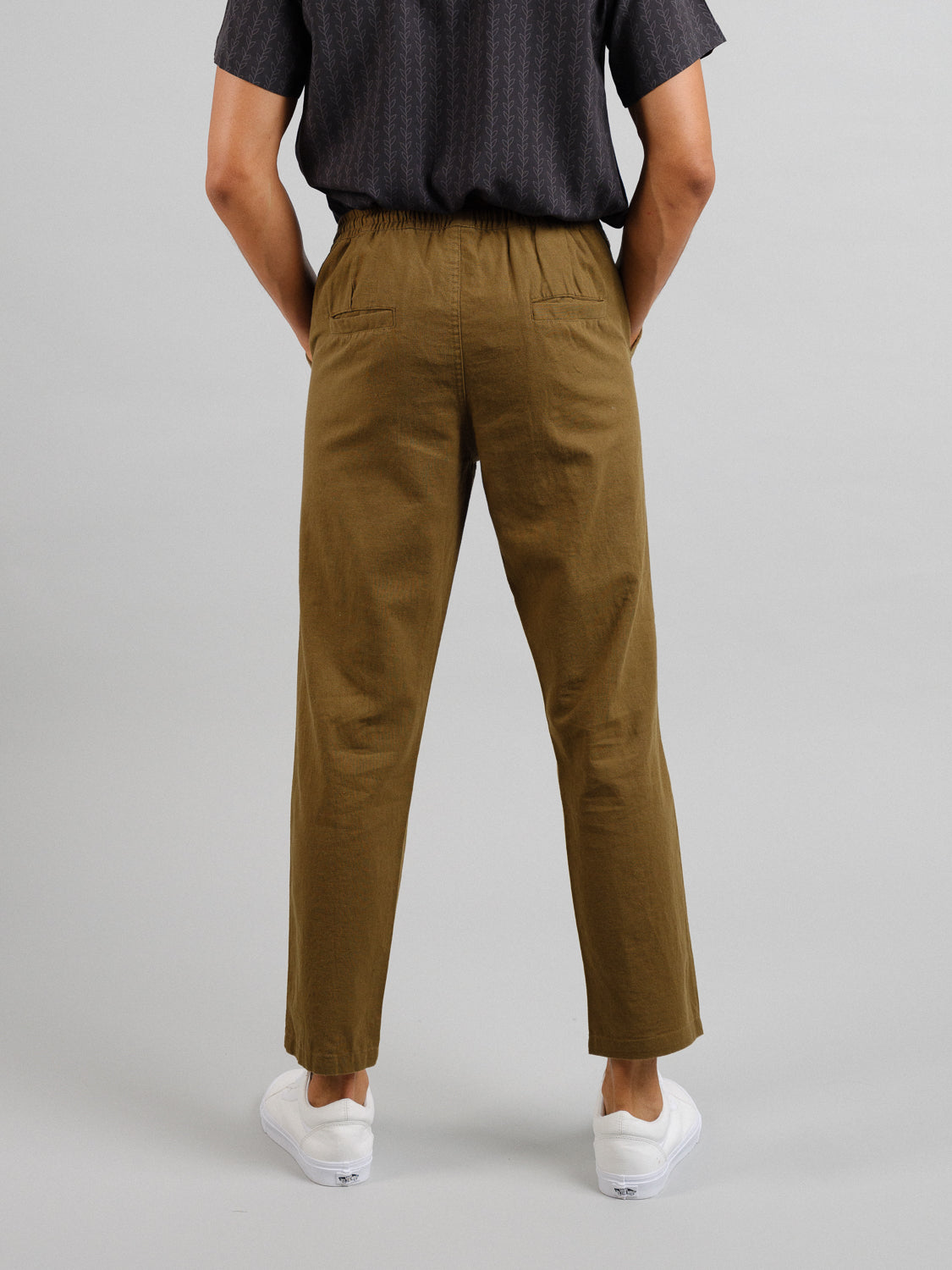 Rear view of a person in ID Menswears Laguna Linen Cotton Drawstring Pant with a relaxed crop fit, coupled with a dark short-sleeve shirt and white slip-on shoes. The casual outfit is set against a plain light gray backdrop.