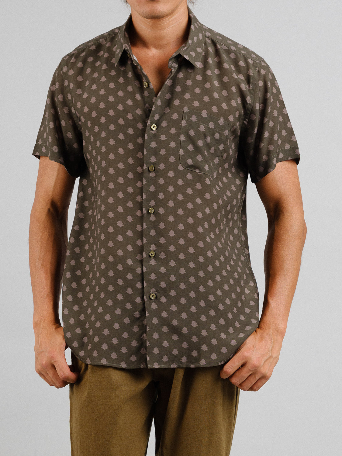 A person models the Ogawara Printed Tencel Linen Shirt from ID Menswear, featuring short sleeves and a light pattern on a dark background. Ideal for summer with a chest pocket, it’s paired with brown pants in a cropped image from mid-thigh up.