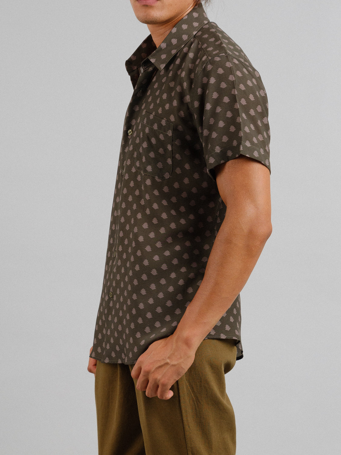 Someone in a short-sleeved, olive green Ogawara Printed Tencel Linen Shirt by ID Menswear stands against a gray background, with hands at their sides and gazing slightly left, showcasing an effortless style ideal for any summer wardrobe.