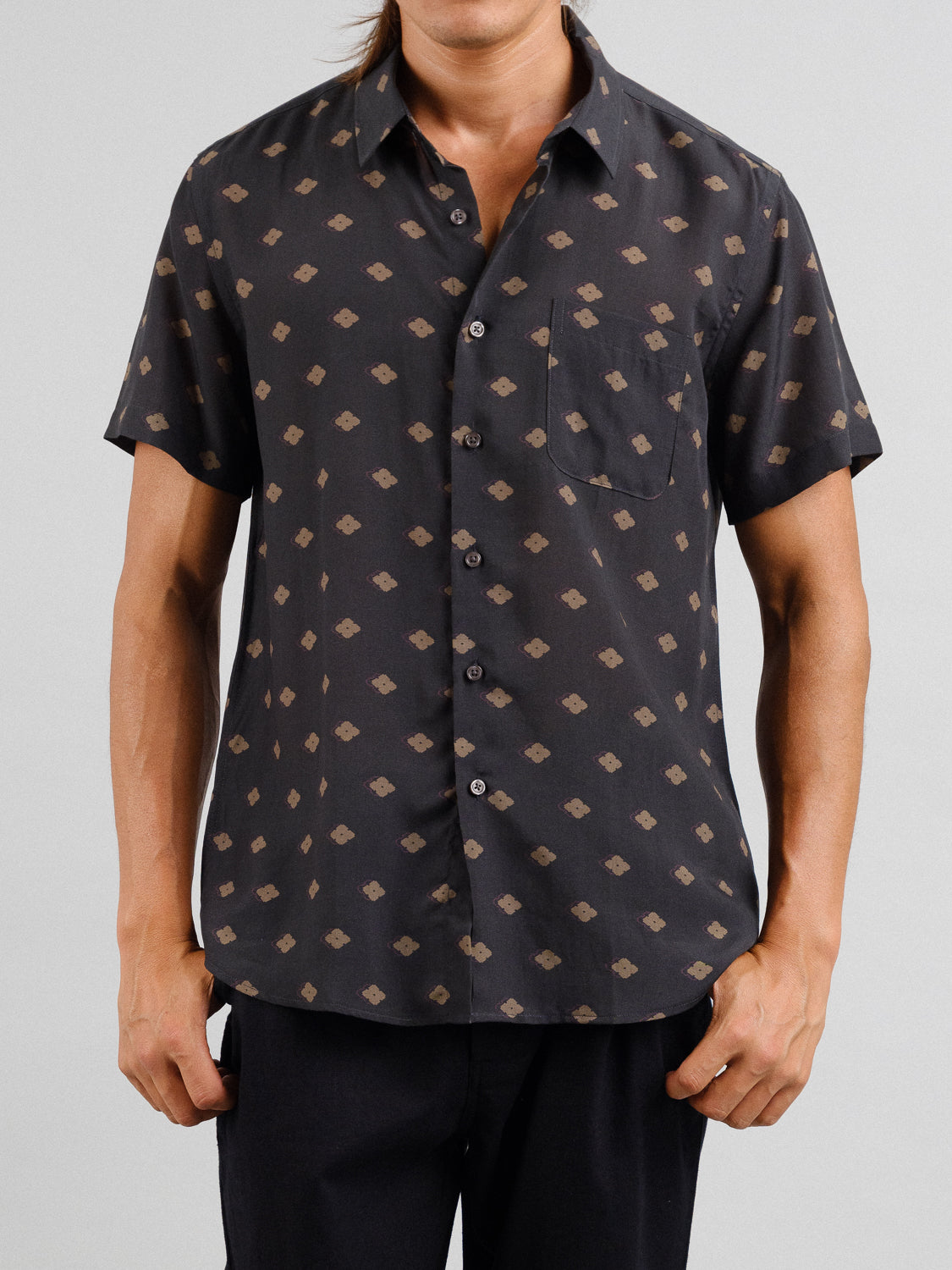 Someone is wearing the short-sleeve Keda Printed Tencel Linen Shirt by ID Menswear, featuring a small geometric pattern in brown and black. It’s perfect for summer with a front pocket and pairs well with black pants against a plain gray backdrop.