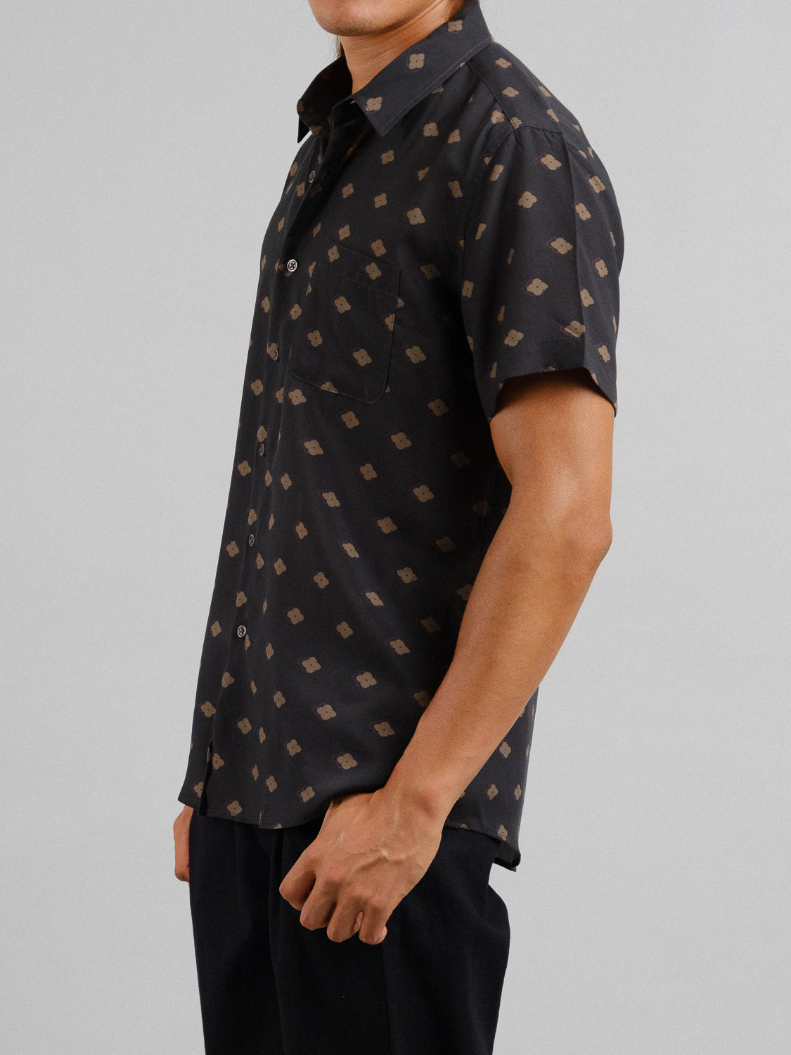 A man in a black ID Menswear Keda Printed Tencel Linen Shirt, ideal for summer with its tan geometric pattern and front pocket, stands against a plain gray background.