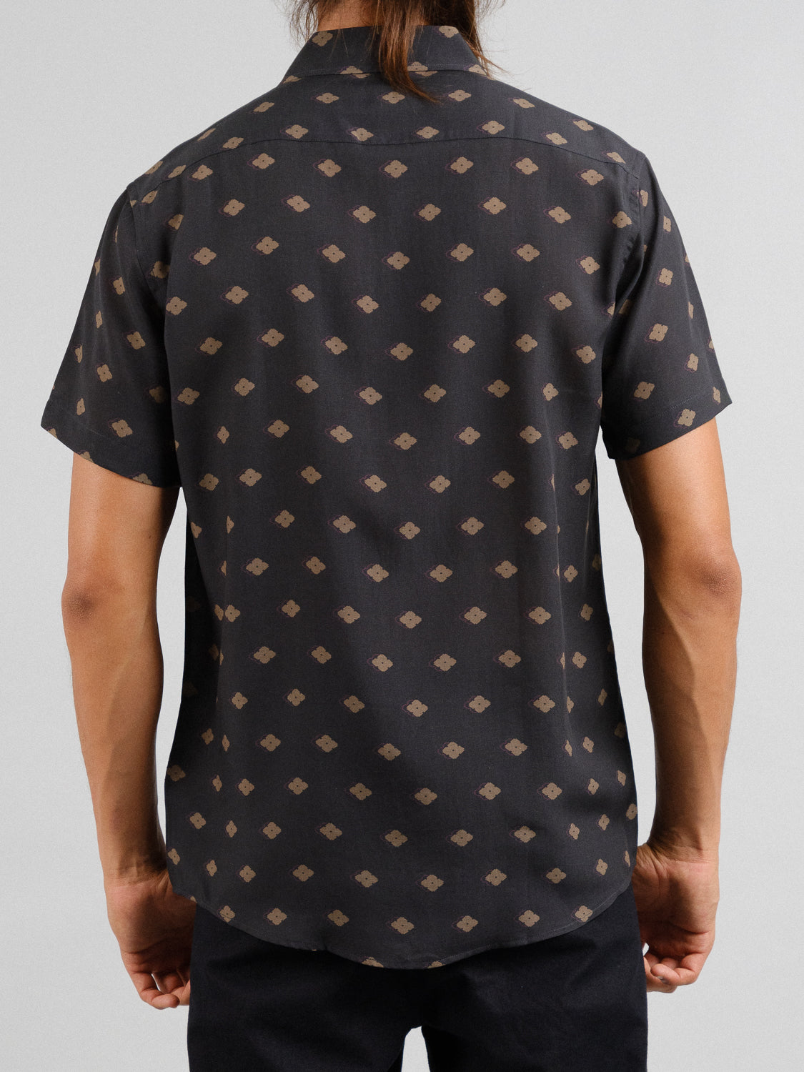 A person with long hair seen from the back wears the Keda Printed Tencel Linen Shirt by ID Menswear—a short-sleeve, black button-up with a geometric brown pattern—paired with black pants against a plain background, making it an ideal summer wardrobe piece.