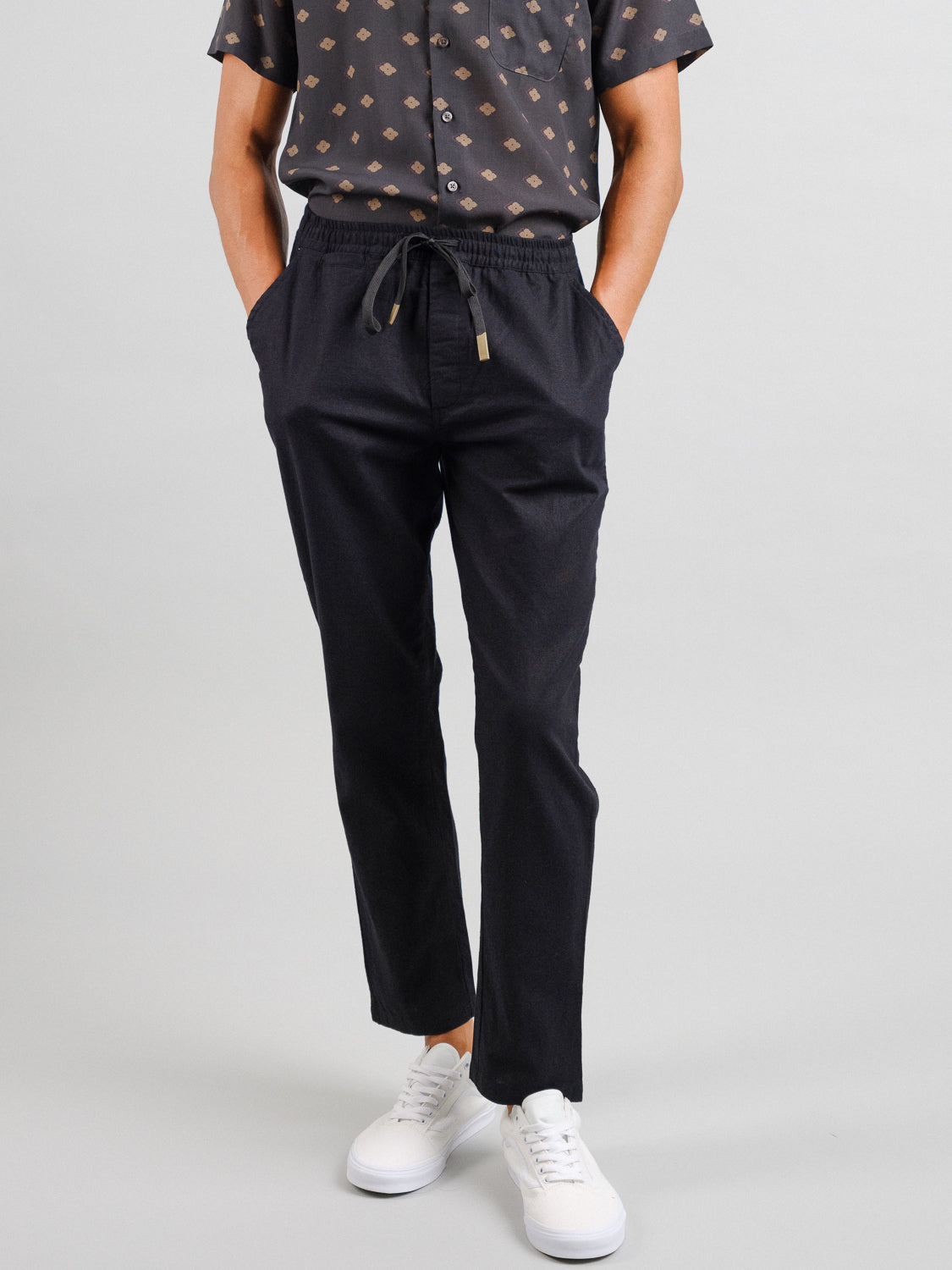 A person wears Laguna Linen Cotton Drawstring Pants in black by ID Menswear, paired with a dark patterned shirt and white sneakers. Hands in pockets, their relaxed crop fit stands out against a light backdrop, highlighting the effortless style.