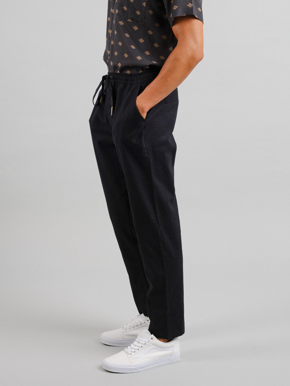 A person wearing a short-sleeved patterned shirt, ID Menswears Laguna Linen Cotton Drawstring Pants in black with a relaxed crop fit, stands with hands in pockets against a gray background, sporting stylish white sneakers.