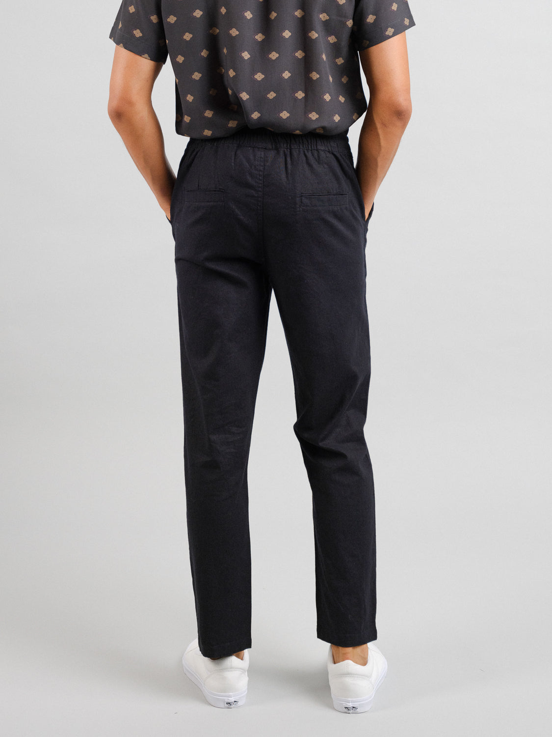 A person is seen from behind wearing ID Menswear Laguna Linen Cotton Drawstring Pants and a dark patterned shirt, hands in pockets. White linen cotton sneakers complete the look, set against a plain backdrop.