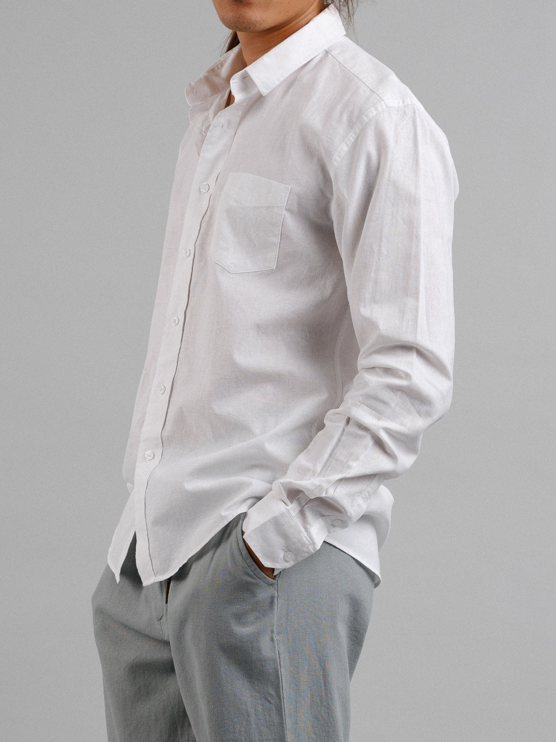 A person models the ID Menswear Goa Linen Cotton Long Sleeve Shirt in white and light gray pants, standing against a plain gray background with hands in pockets.