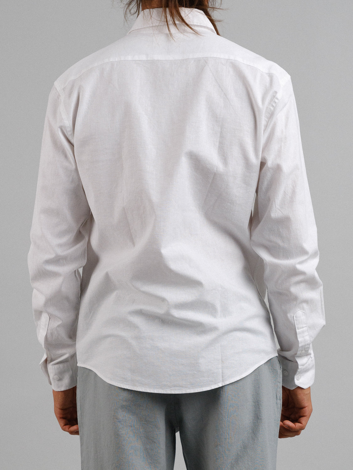 A person with long hair stands with their back to the camera, wearing an ID Menswear Goa Linen Cotton Long Sleeve Shirt in white and light gray pants against a plain gray background.