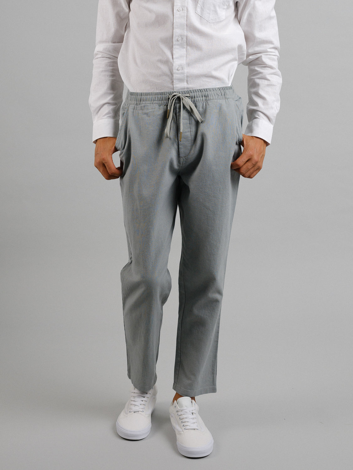 A person stands against a plain gray background, wearing a white long-sleeve shirt, white sneakers, and light gray Laguna Linen Cotton Drawstring Pants by ID Menswear with a relaxed crop fit. Their hands are in the pockets of the pants.