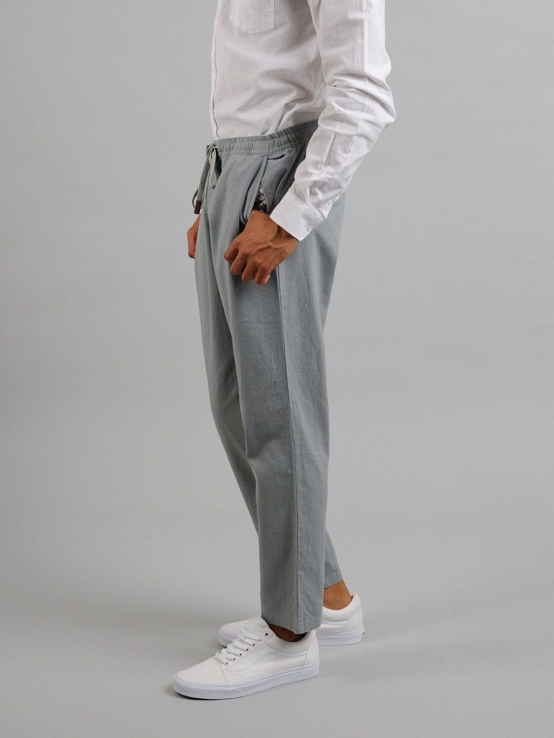 A person in an ID Menswear white long-sleeve shirt, light gray Laguna Linen Cotton Drawstring Pant with a relaxed crop fit, and white sneakers stands against a gray background, facing sideways to the left.