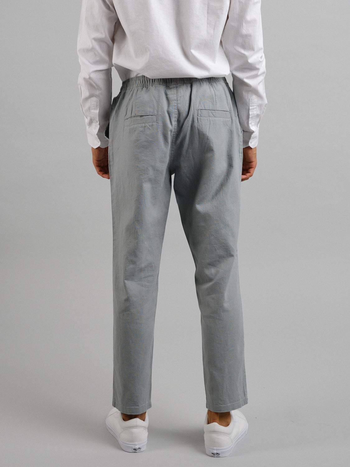 A person is shown from the back wearing a white long-sleeve shirt, white sneakers, and ID Menswears Laguna Linen Cotton Drawstring Pant in light grey with a relaxed crop fit. The pants have an elastic waistband and two back pockets against a plain background.
