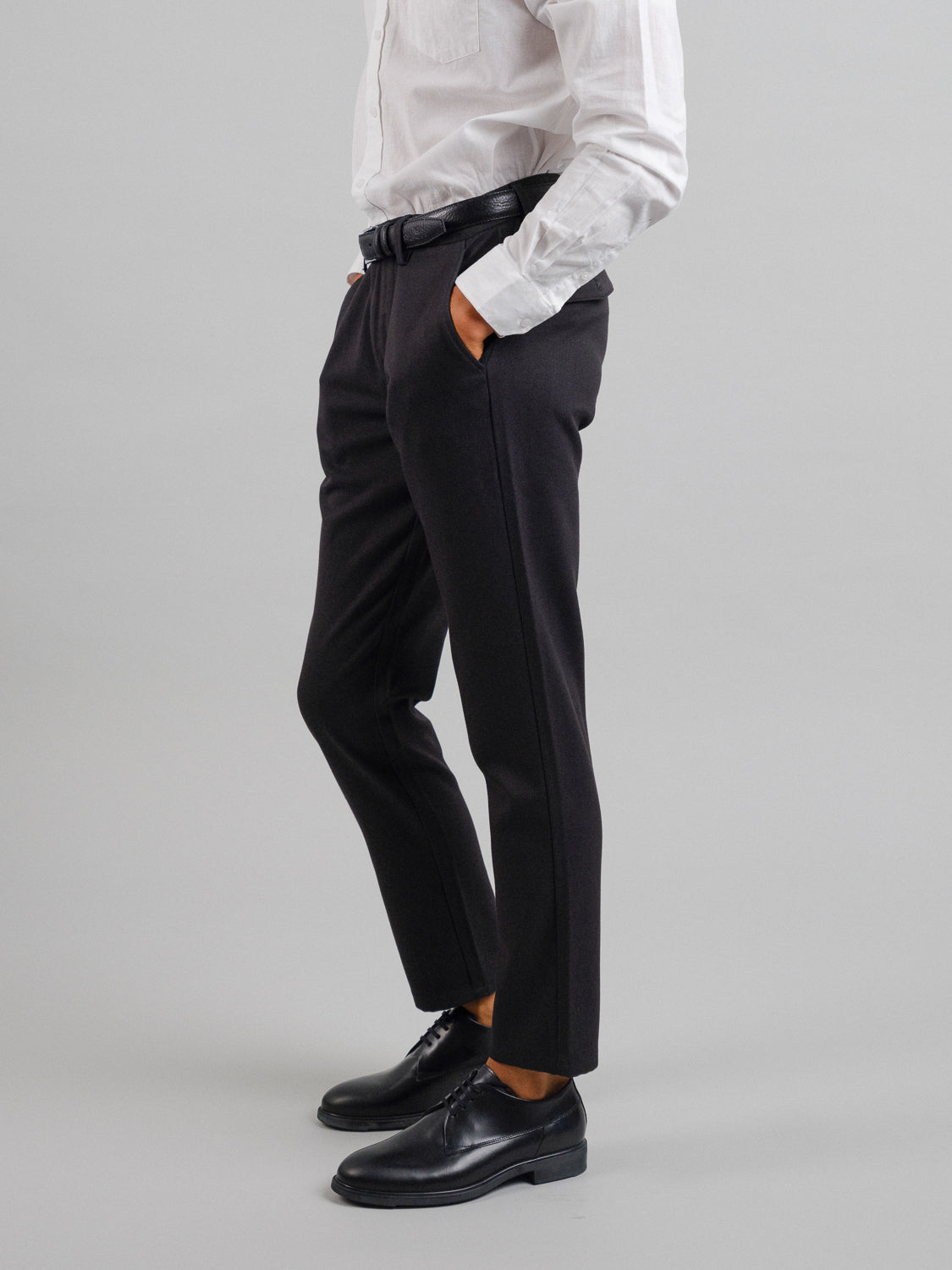 A person exudes timeless style in a white long-sleeve shirt tucked into ID Menswears Kensington Wool Blend Chino and black tailored pants, hands in pockets. Completed with a black belt and dress shoes, this ensemble stands perfectly against a plain gray background.