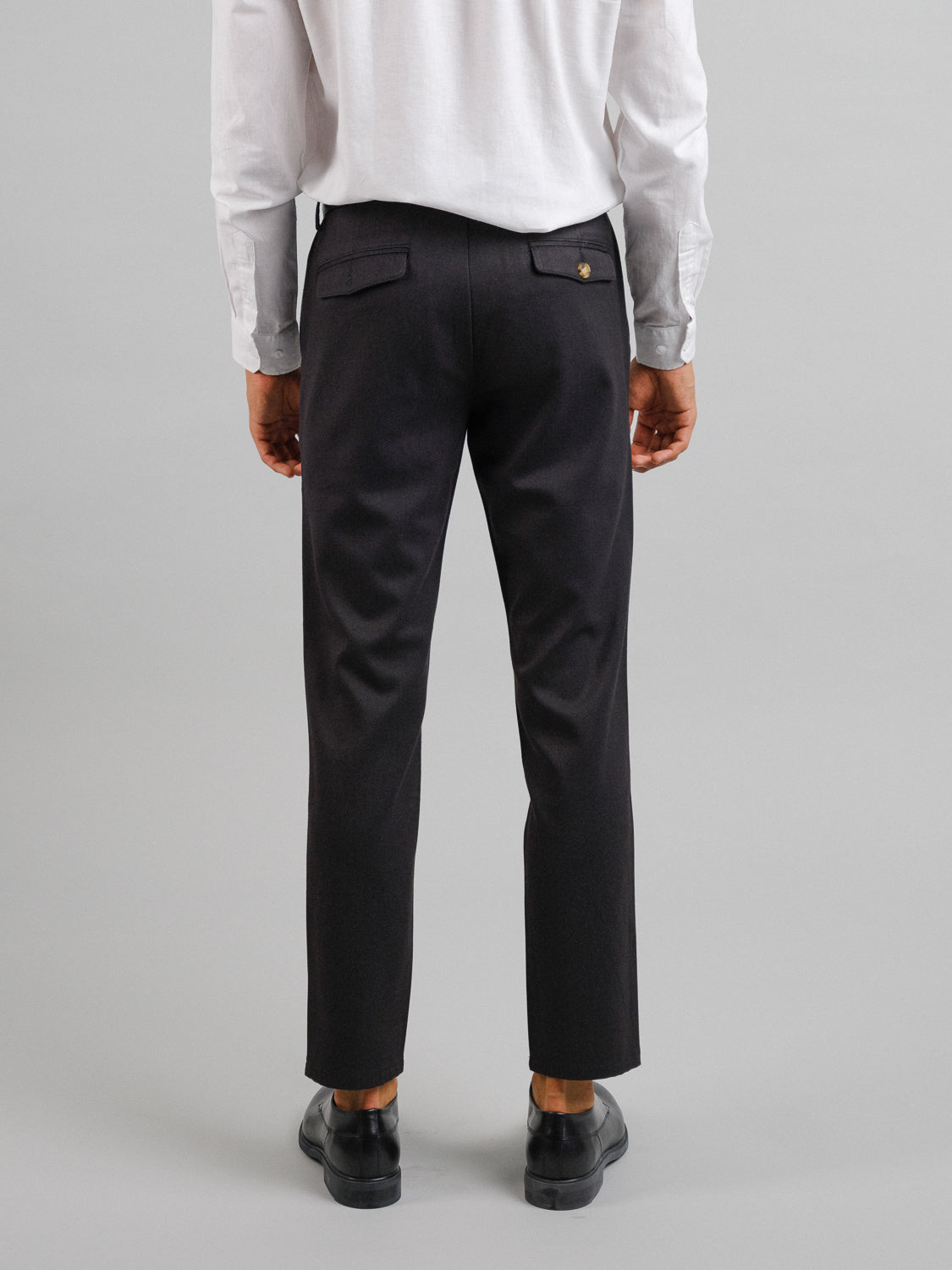 Back view of a person wearing a white long-sleeve shirt and black Kensington Wool Blend Chinos by ID Menswear. Standing against a neutral background, their black shoes add formality to this timeless style, making it suitable for any occasion.