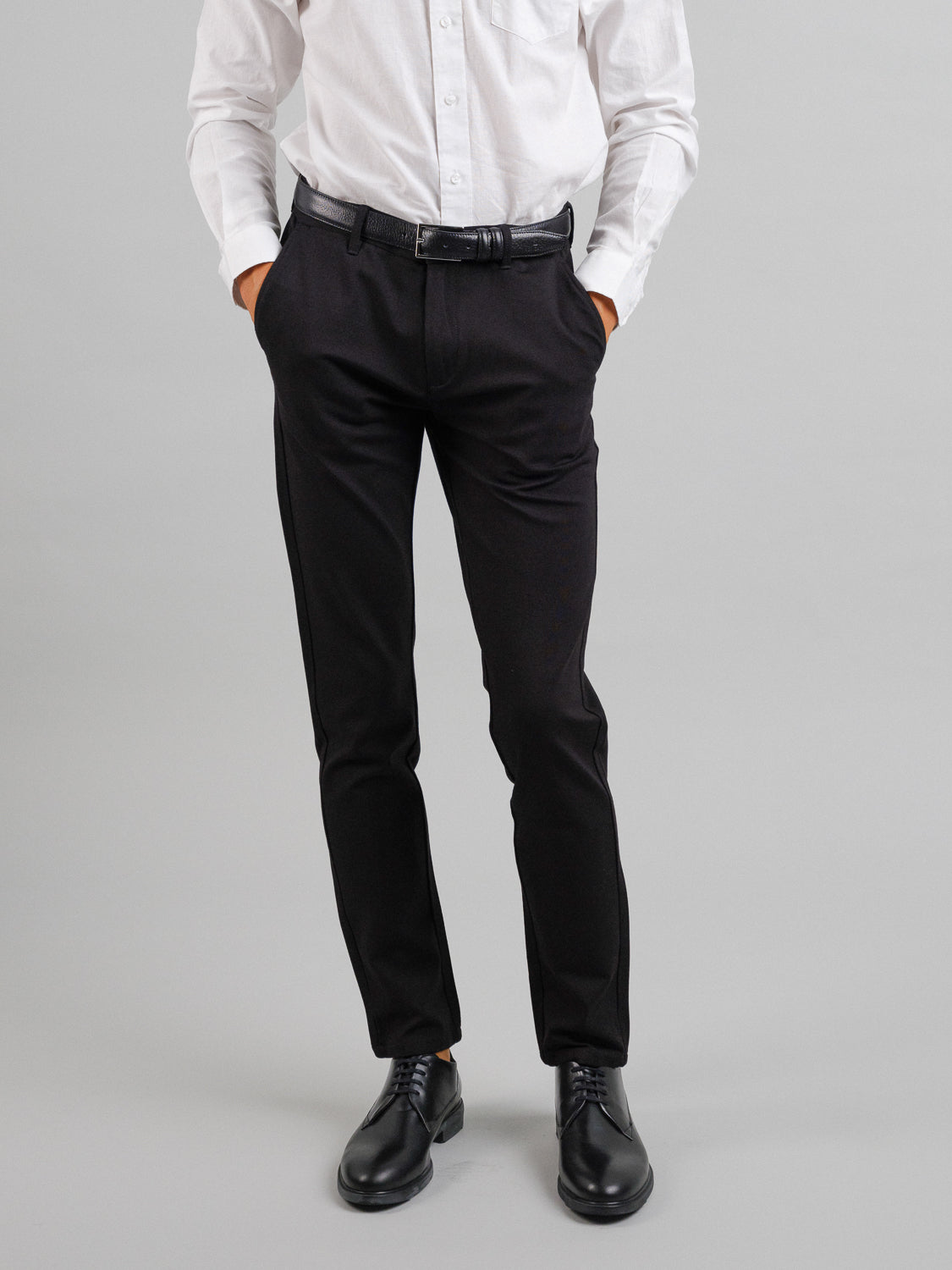A person stands with hands in pockets against a gray background, wearing a slim-fit white dress shirt, black belt, black dress shoes, and ID Menswear’s Montrose 4-Way Stretch Dress Pant. Their head is not visible, creating a business casual look.