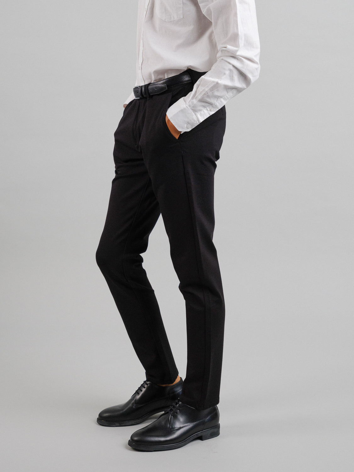 A person stands with hands in pockets, wearing a slim-fit white long-sleeve shirt, a black belt, black dress shoes, and ID Menswear's Montrose 4-Way Stretch Dress Pant, against a plain light gray background. The upper part of the head is not visible.