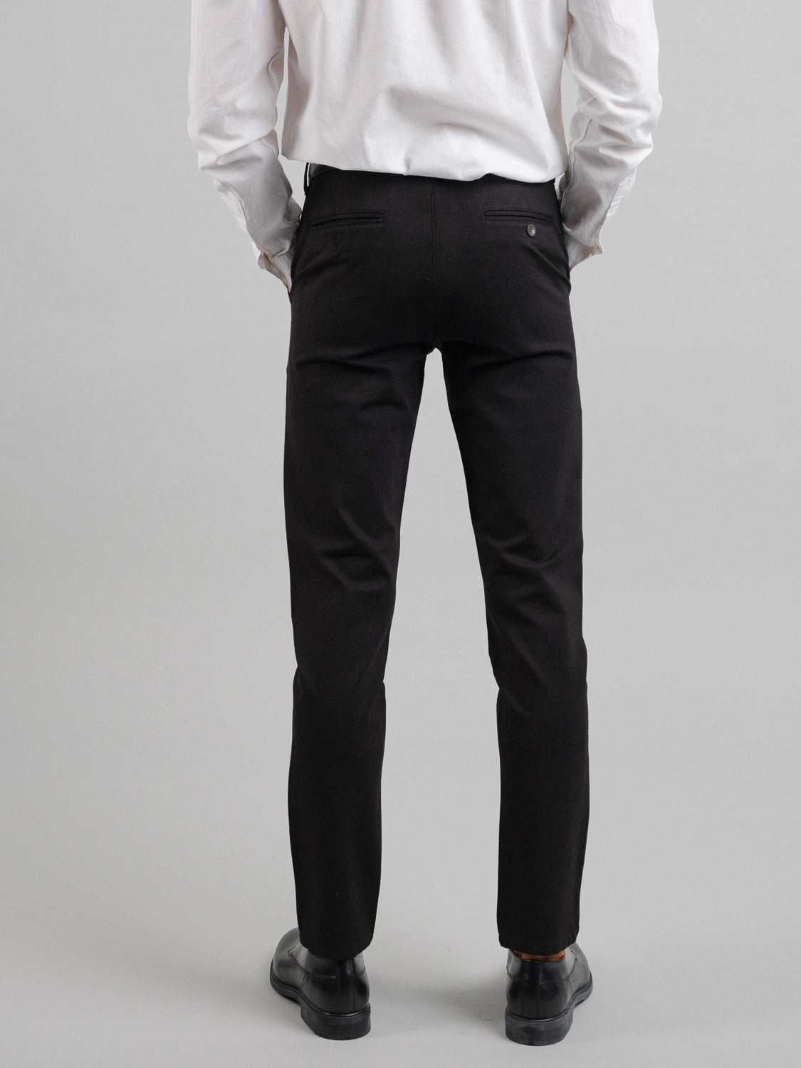 A person is seen from behind wearing ID Menswear’s Montrose 4-Way Stretch Dress Pant in black, paired with a white long-sleeve shirt and black shoes, against a plain gray background. Only their lower back, legs, and parts of their arms are visible.