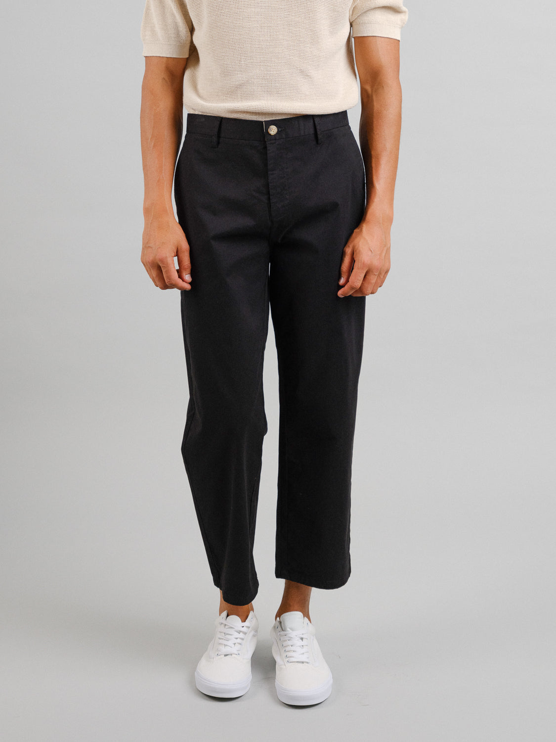 A person stands against a gray background wearing black Bigboy Relaxed Flooded Chino pants by ID Menswear, paired with a short-sleeve beige shirt and white sneakers. The relaxed, baggy fit of the chinos enhances the casual look.