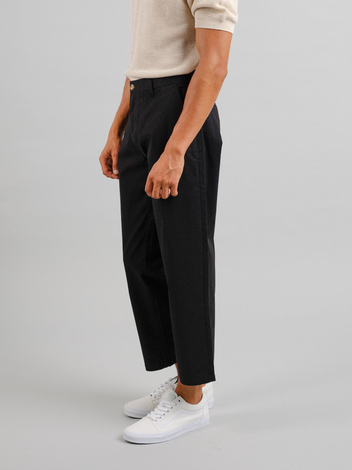 A person wears a beige short-sleeve shirt, ID Menswears Bigboy Relaxed Flooded Chino in black for a baggy fit, and white sneakers. Against a light gray backdrop, their hands rest at their sides, embodying lightweight flexibility.