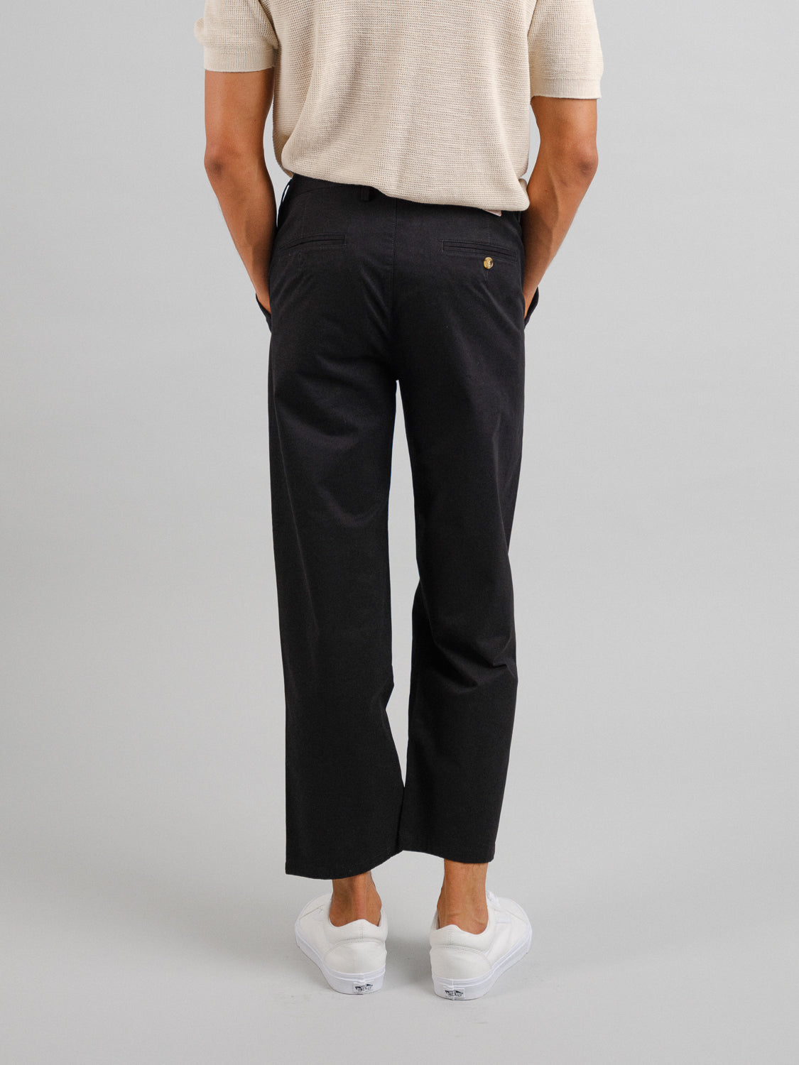 Facing away from the camera, a person wears a light short-sleeve shirt, white sneakers, and ID Menswears Bigboy Relaxed Flooded Chino with a dark hue, relaxed fit, and baggy style against a plain gray backdrop.