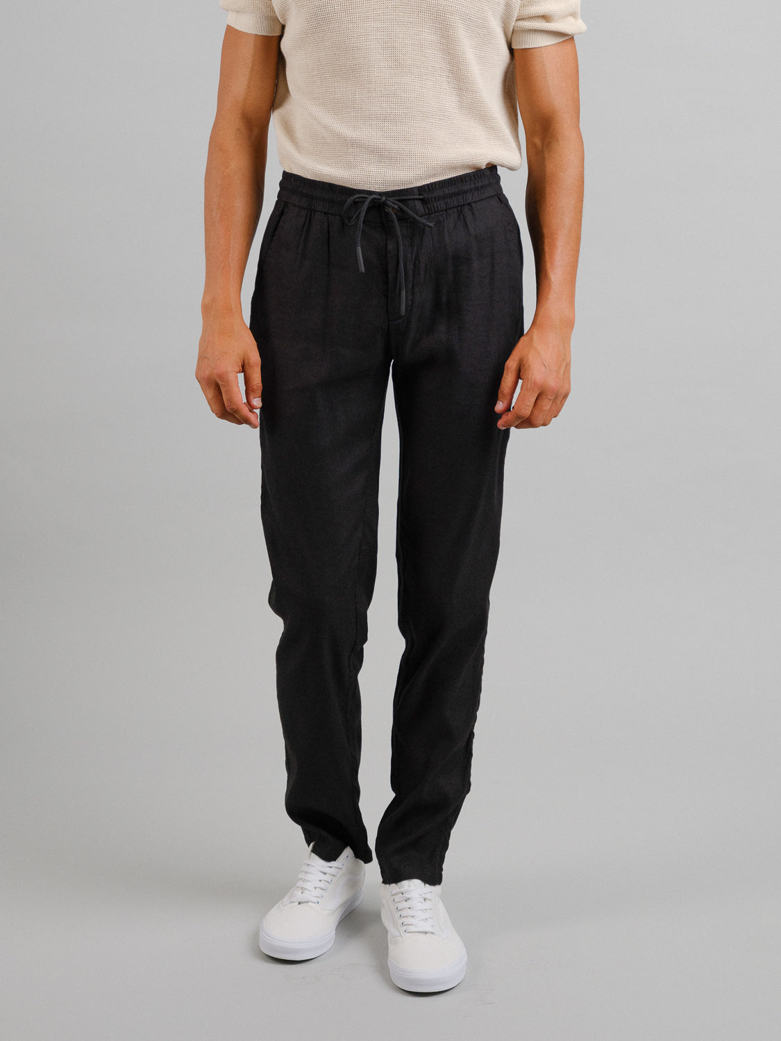 A person stands against a gray background wearing a cream short-sleeve shirt, white sneakers, and comfortable Bedford23 Linen Blend Drawstring Pant by ID Menswear, featuring an elastic waistband. The focus is on this relaxed outfit from neck to shoes.