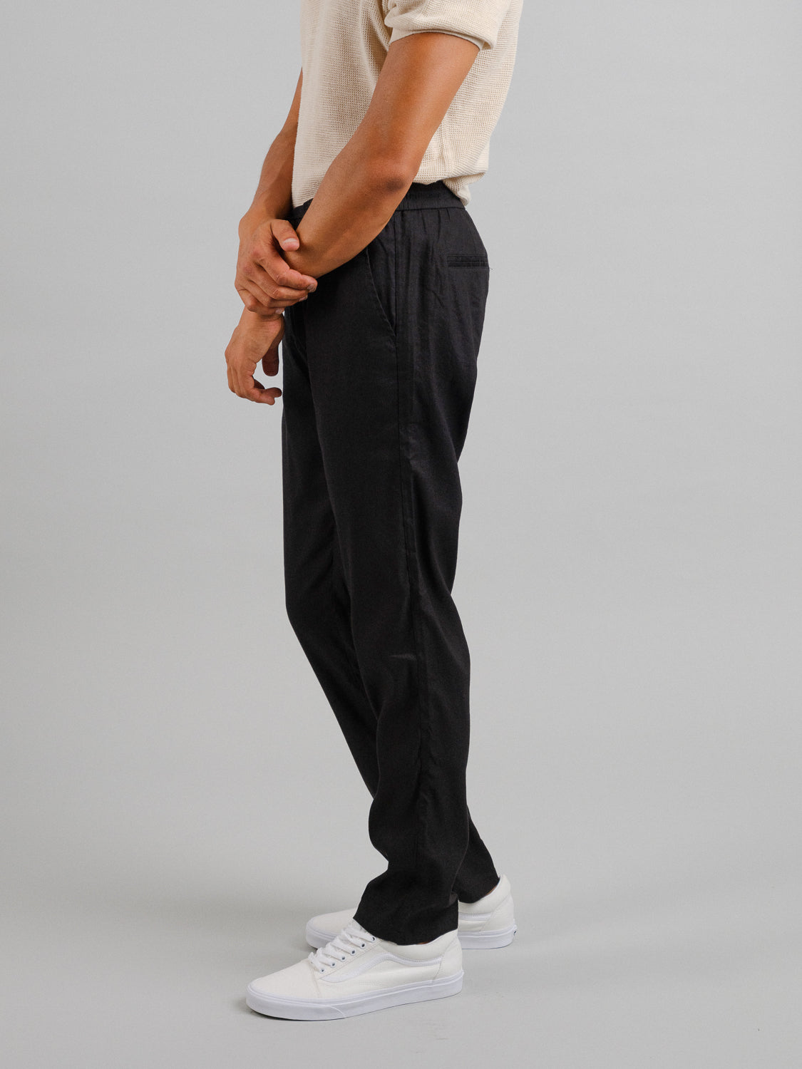 The person stands sideways against a plain backdrop, wearing a beige shirt and Bedford23 Linen Blend Drawstring Pant by ID Menswear. The pants have an elastic drawstring waistband and are paired with white sneakers, with their arms lightly crossed in front.