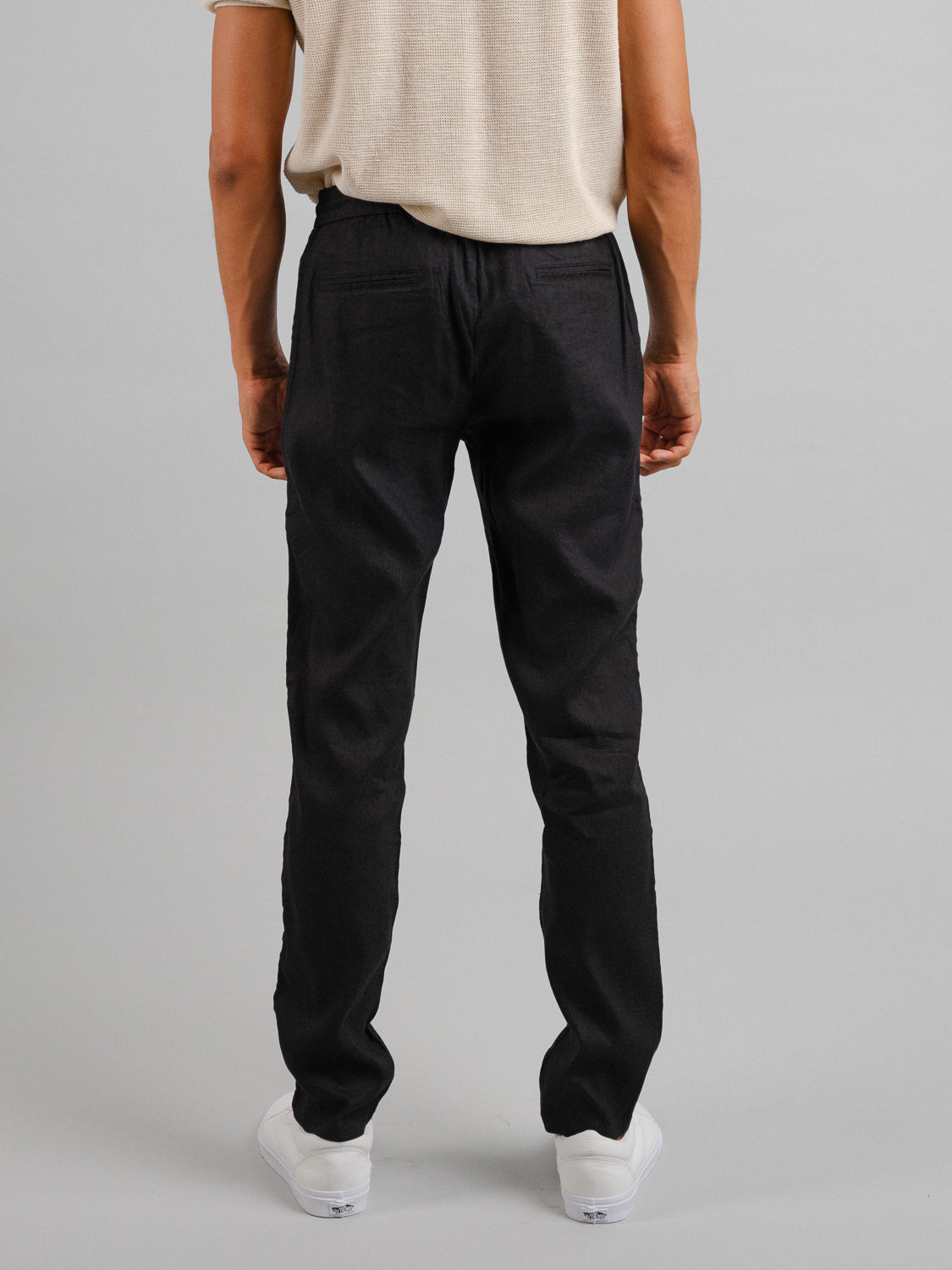 The person stands back to the camera in a beige shirt, ID Menswears Bedford23 Linen Blend Drawstring Pant with an elastic waistband, and white sneakers against a plain grey background.