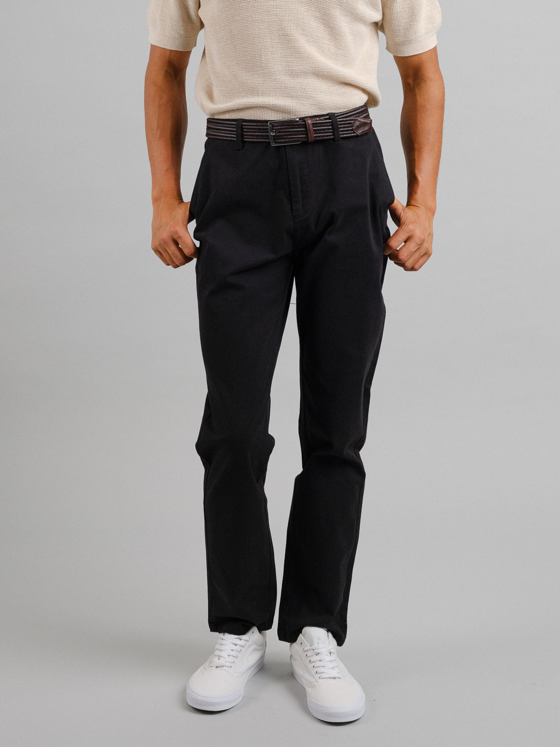 A person sports a light beige short-sleeve shirt with ID Menswears Bushwick Twill Stretch Chino, relaxed-fit black trousers. Completing the look with a belt and white sneakers, they stand against a plain gray background, hands on pants exuding casual charm.