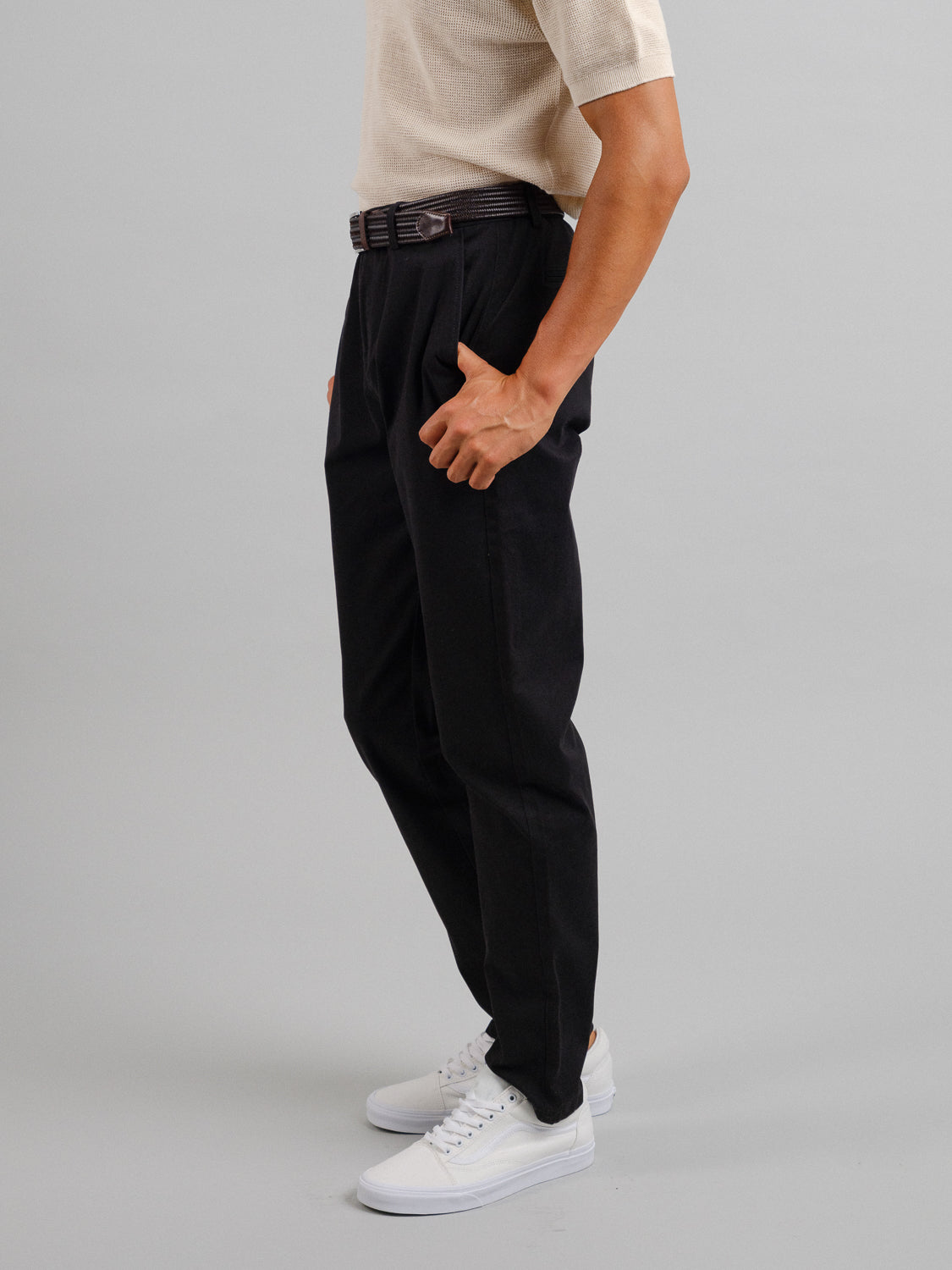 A person in a beige short-sleeve shirt and ID Menswears Bushwick Twill Stretch Chino stands in profile against a gray background, with white sneakers and their right hand tucked into the pocket of the black chinos.