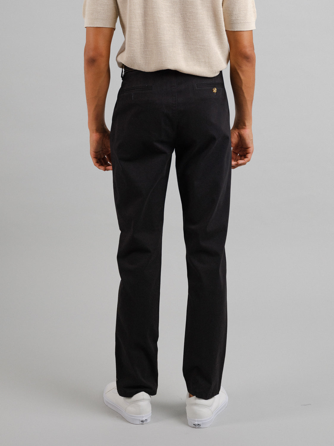 With their back to the camera, a person wears a short-sleeved, light shirt and relaxed-fit Bushwick Twill Stretch Chino by ID Menswear. The stylish white shoes complete the look against a plain gray backdrop.