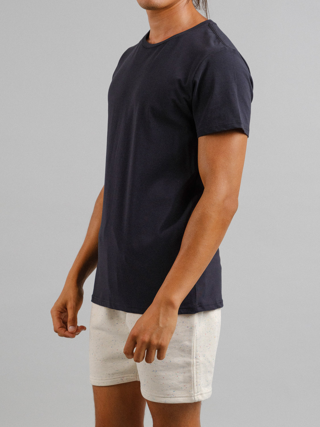 A person wears a black Form Crew Neck Tee by ID Menswear and light shorts against a gray backdrop, standing at an angle with relaxed arms. The tees form-fitting design and comfy fabric highlight their effortlessly stylish, casual look.