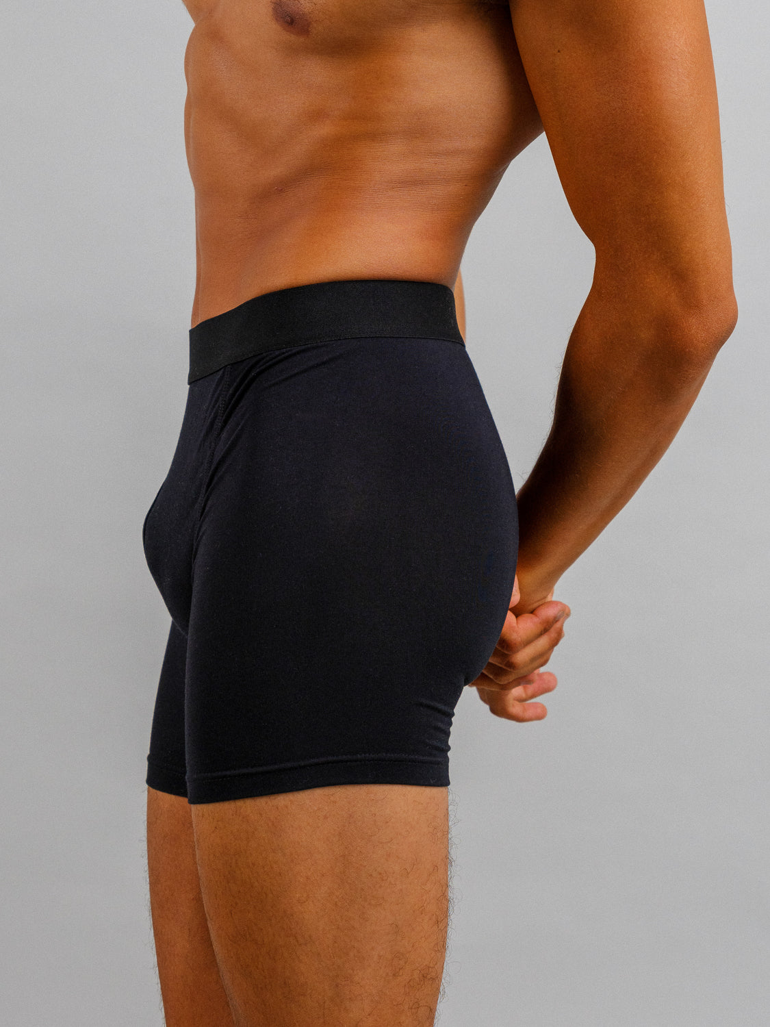 A shirtless person wearing ID Menswears black modal boxer briefs from the ID Modal Boxer Briefs 3 Pack stands sideways with hands behind their back against a neutral gray background, highlighting the stretch and everyday comfort of the garment.