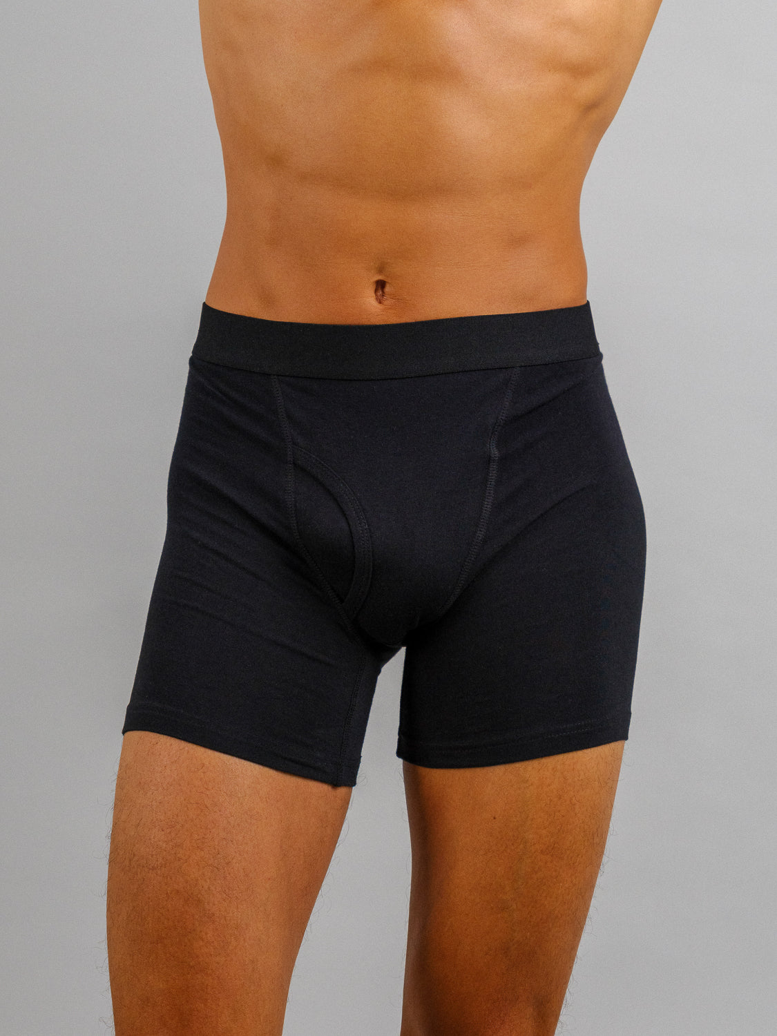 A person wears ID Menswears black boxer briefs from the Organic Cotton Boxer Briefs 3 Pack, made from stretchable organic cotton. They stand against a plain gray background, focusing on the torso and legs without showing their head.