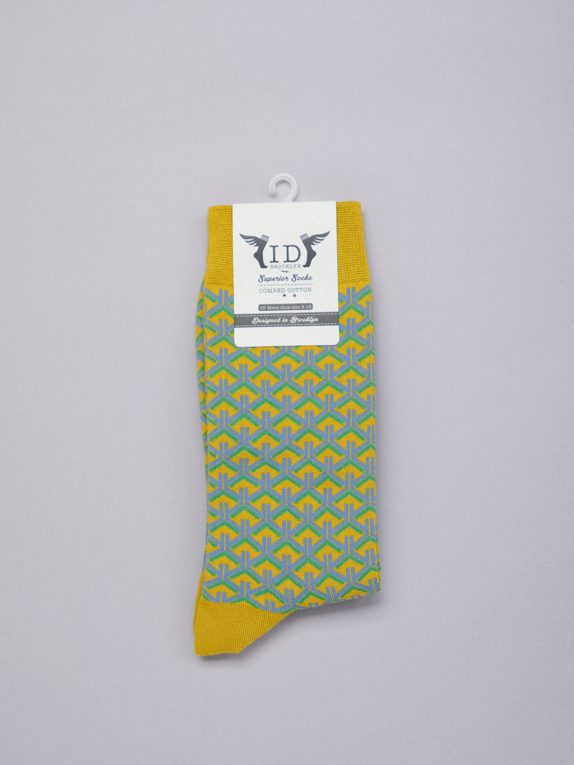 The Wagara Graphic Sock from ID Menswear, featuring a geometric blue and yellow pattern with a yellow cuff and toe, is displayed solo against a plain backdrop. A white paper label with decorative text and the brand logo adorns the sock.