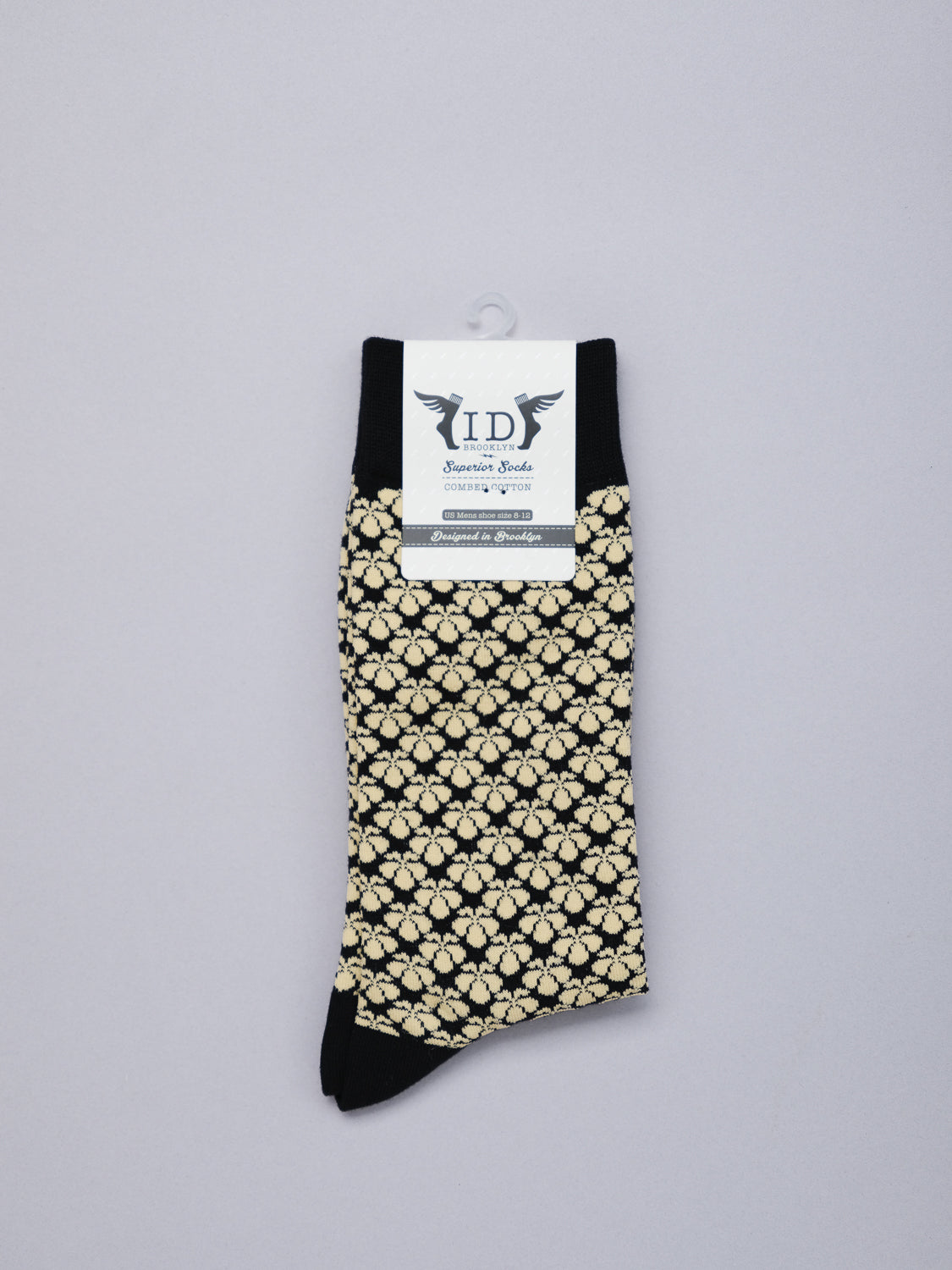 A pair of Toyo Graphic Socks by ID Menswear, featuring a cream floral jacquard pattern on black combed cotton, is displayed against a light gray background. A white label with text and decorative elements highlights its Japanese artistry.