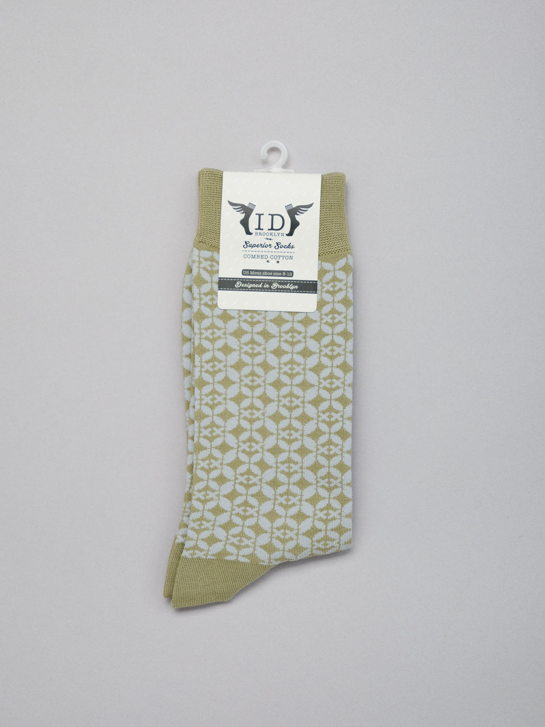 A single Shippo Graphic Sock in beige from ID Menswear is displayed. Made of premium combed cotton, it promises comfort and style. It stands vertically with a branded tag displaying black text and wings, set against a plain light gray background.