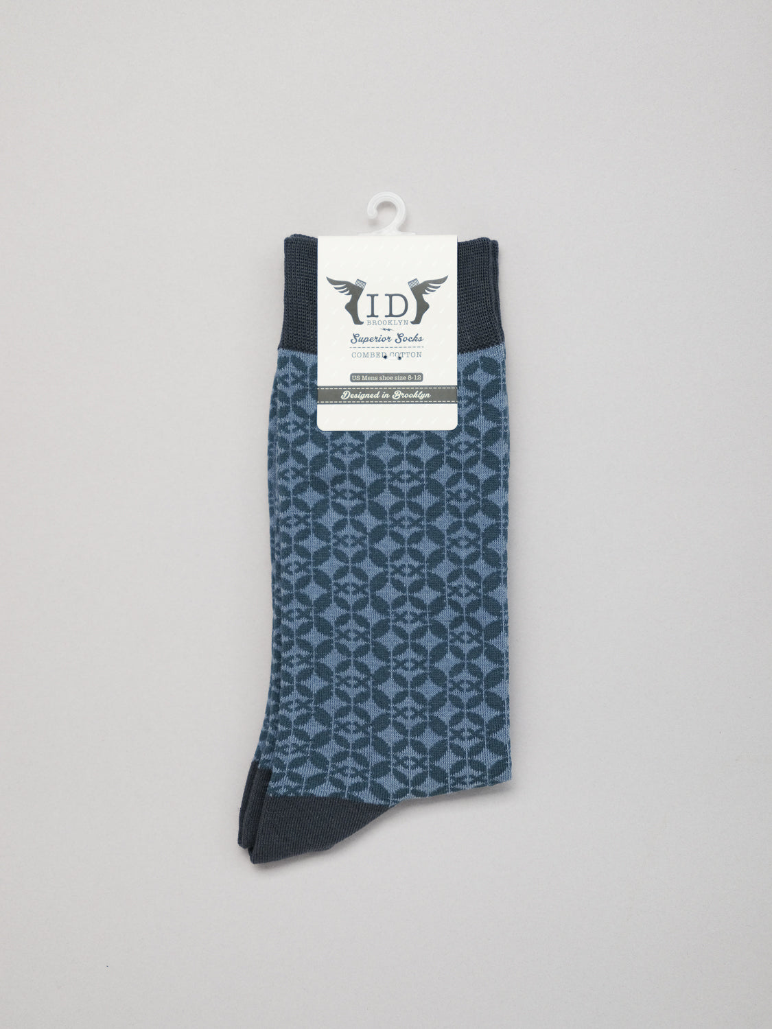 A pair of Shippo Graphic Socks from ID Menswear, featuring a dark blue geometric pattern on a light gray background, hangs prominently. Made of premium combed cotton for comfort, they display a white label with the ID logo and decorative wings.