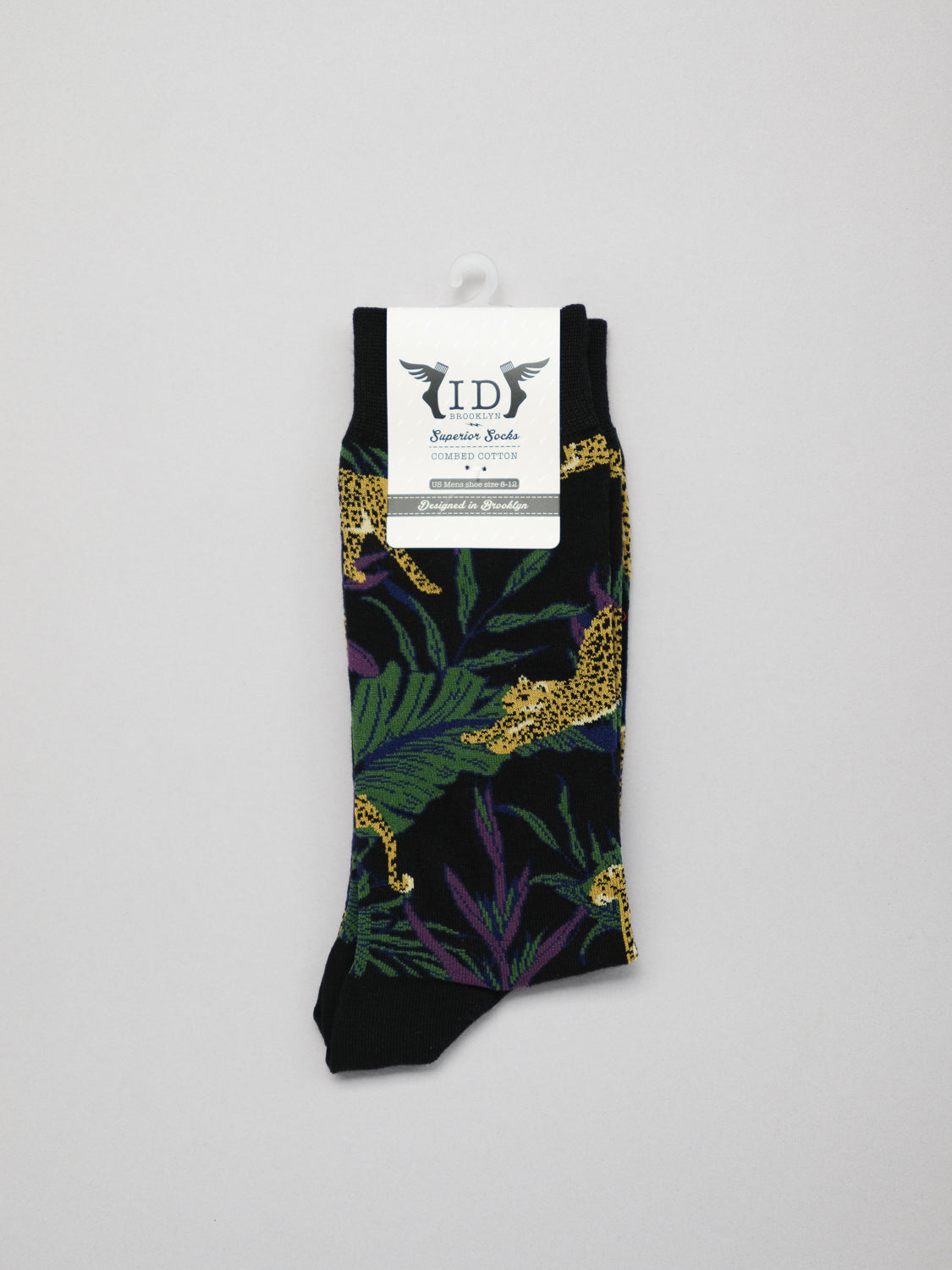 The Jungle Graphic Sock from ID Menswear is a single black sock with vibrant green leaves and yellow cheetahs. Made of combed cotton, it exudes comfort and style. Displayed on a plain gray background, it features a white label with text and logo.