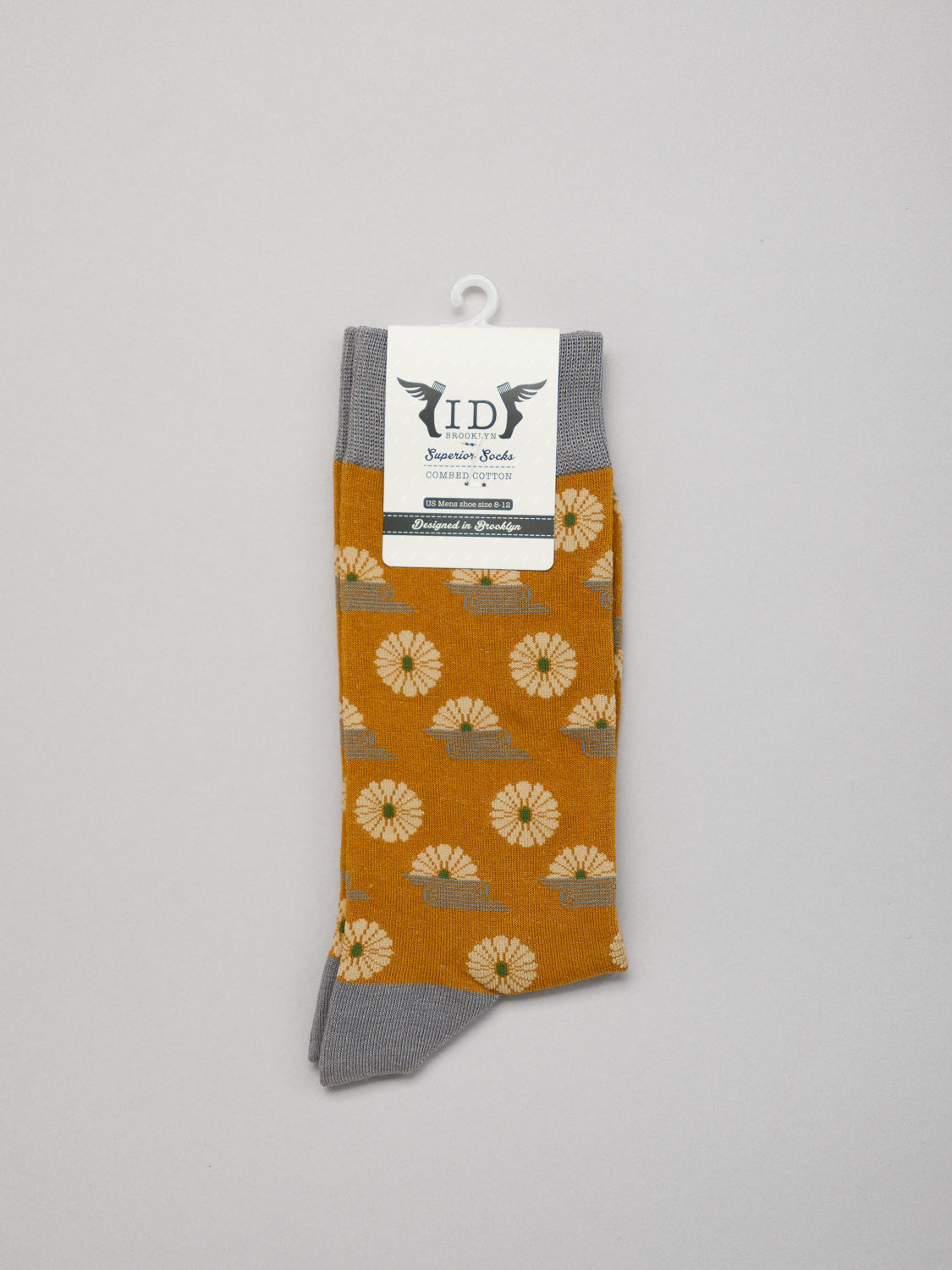 Kiku Graphic Socks by ID Menswear, featuring sunflower patterns on orange combed cotton with gray accents, are displayed on a hanger with a branded label. The backdrop is plain and light gray.