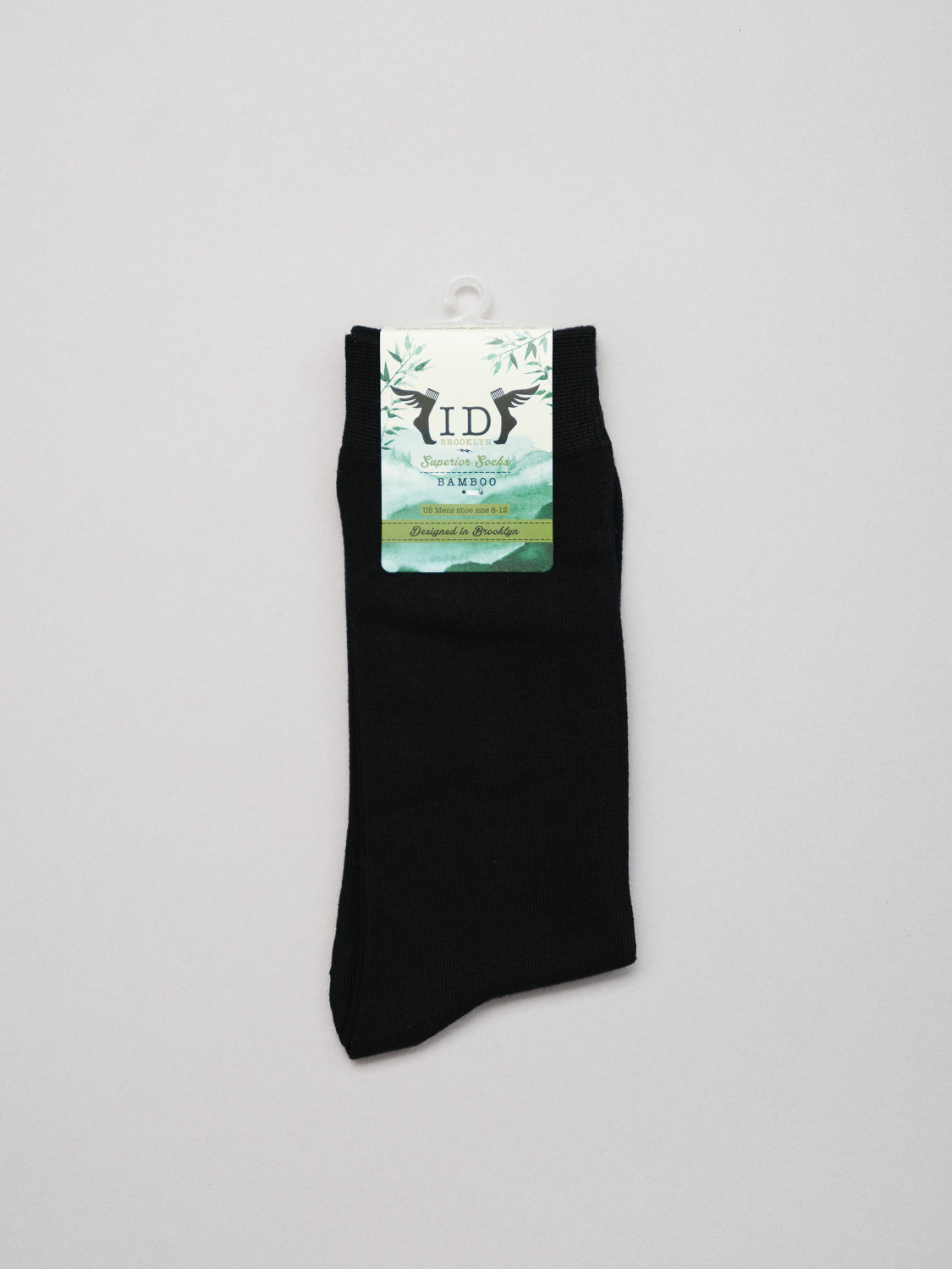 A pair of Cylinder Knit Bamboo Solid Socks from ID Menswear features a black design with a white label that says ID. Made in Japan, the socks are part of the Feminine Touch collection, ensuring breathability. The tag with green leaves underscores sustainability against a plain white background.